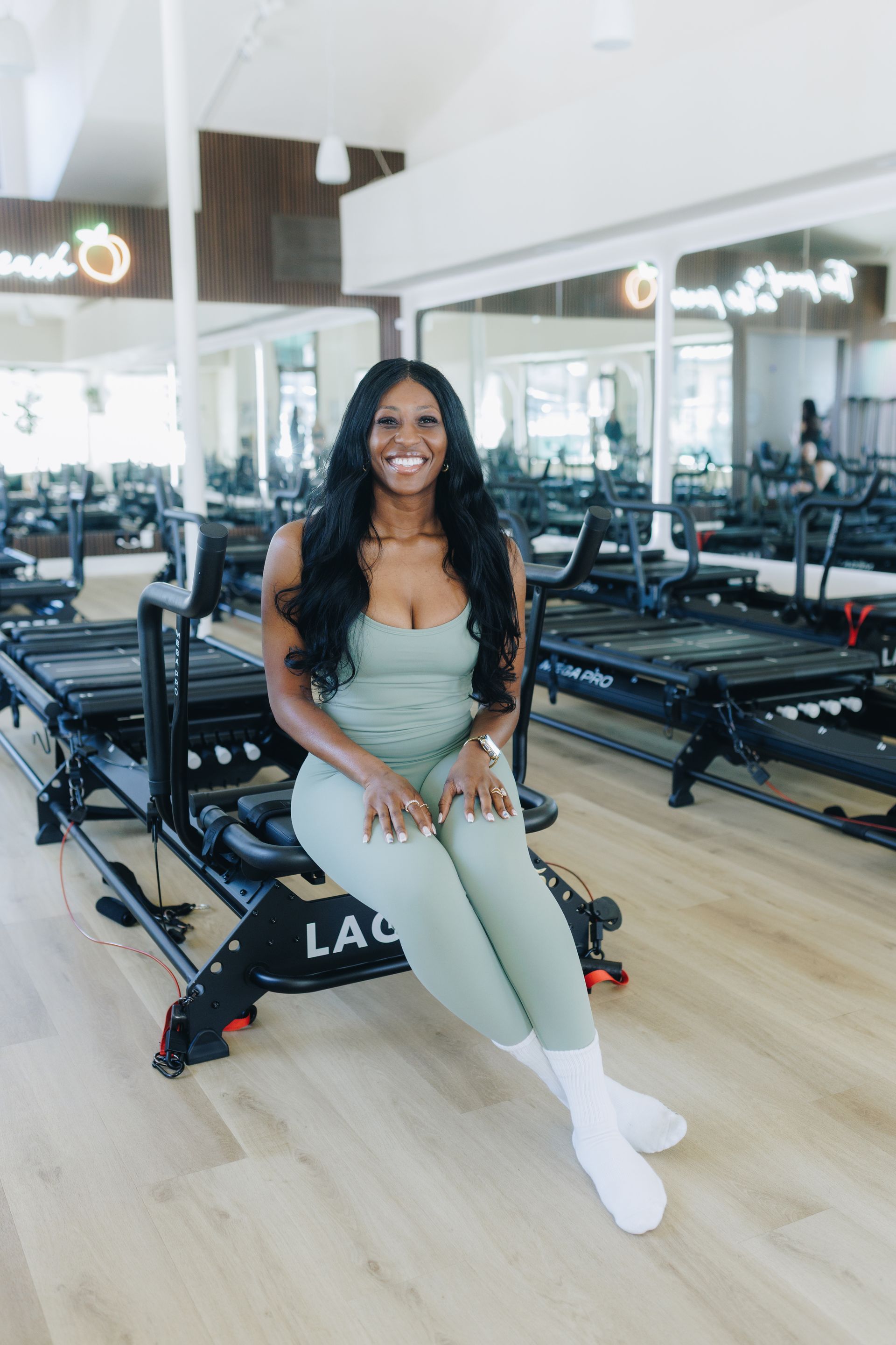 Woman seated on a reformer in a bright Pilates studio, smiling at the camera.