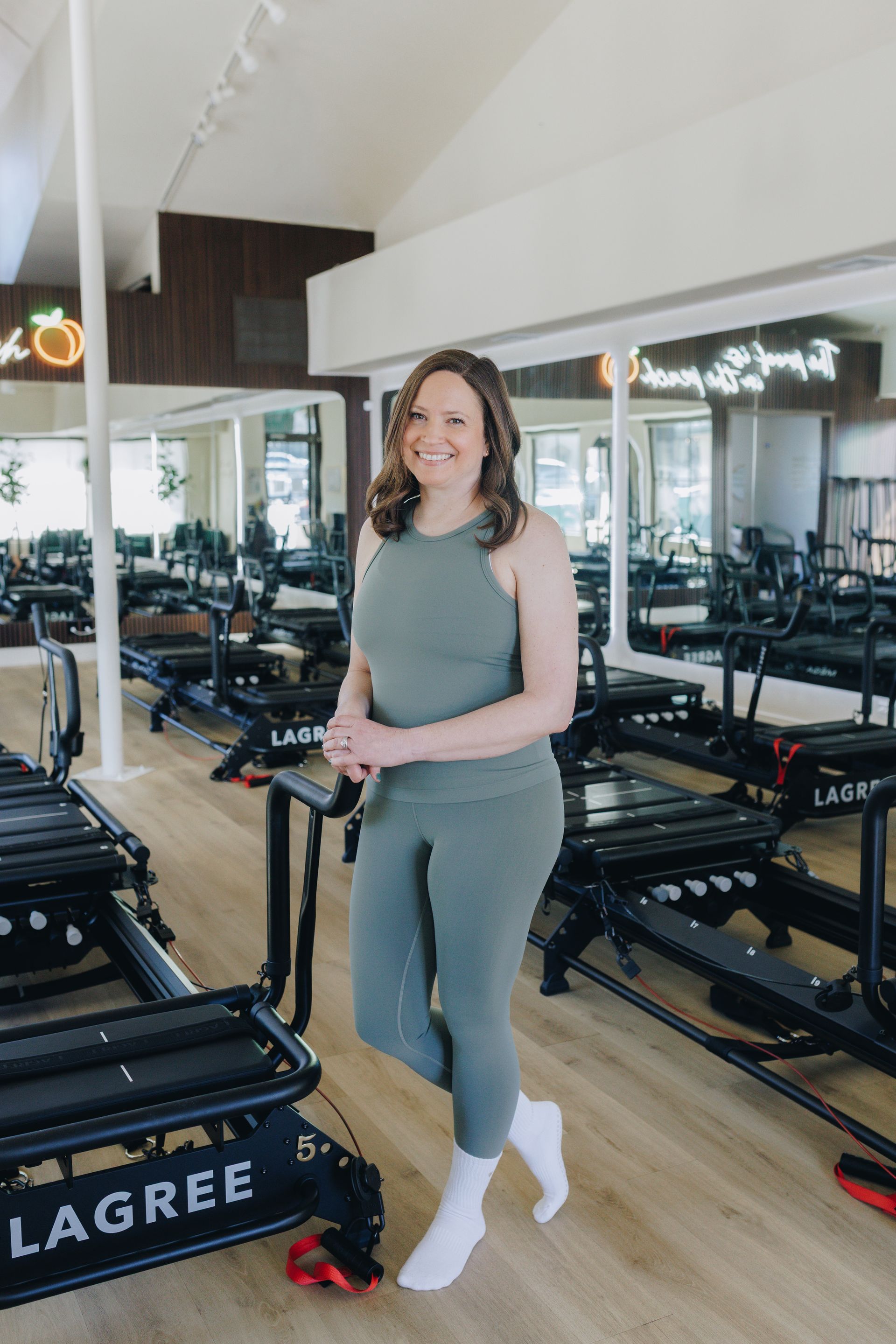 Woman in gray workout clothes standing beside Lagree reformer machines in a mirrored gym