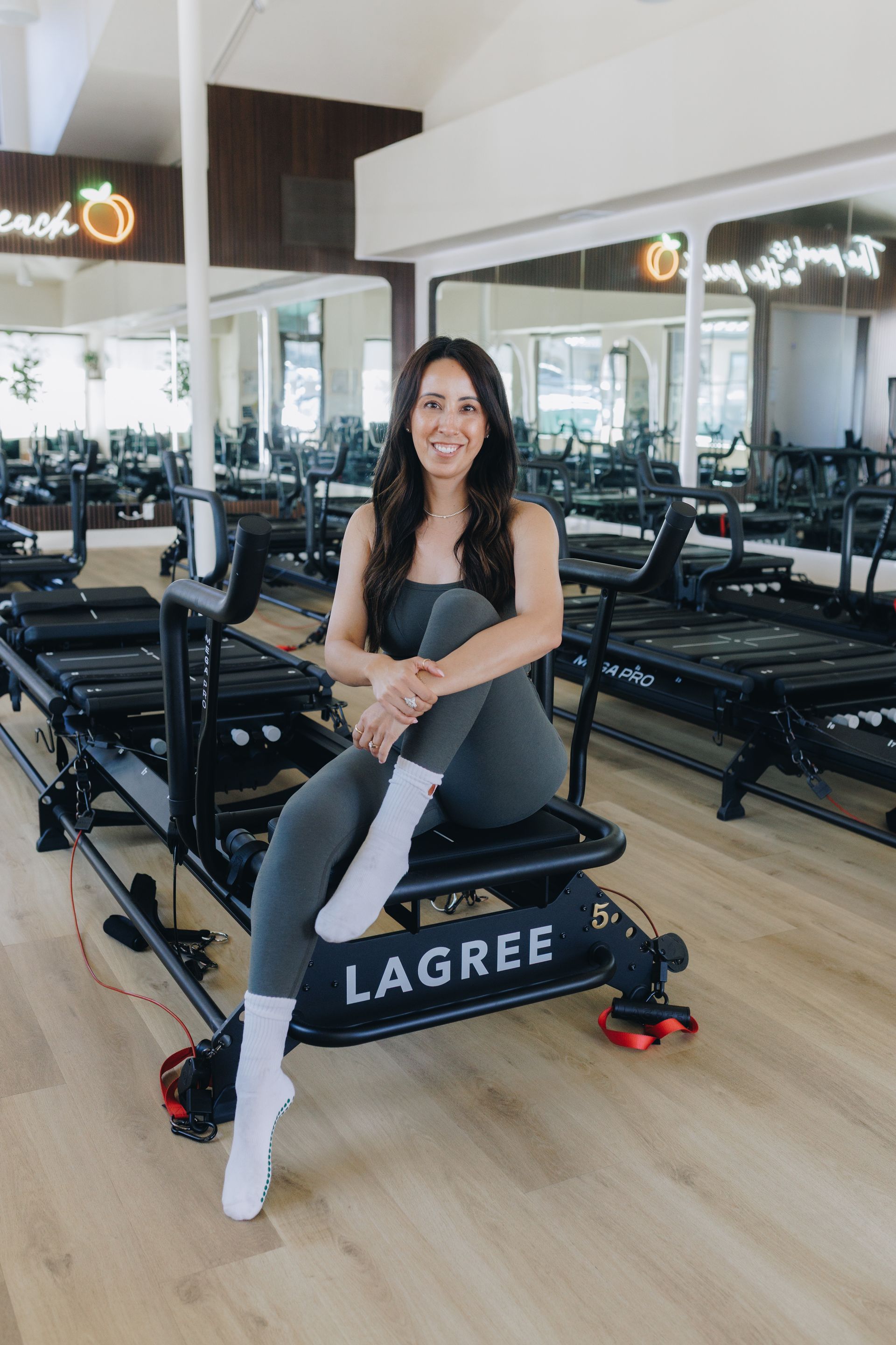 Woman in a gym leaning on a Lagree reformer, smiling, with mirrored walls and wooden floors.