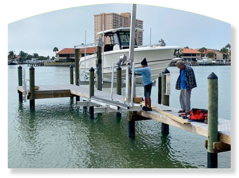 Boat being lifted by a lift at a dock. Two people work on the lift. Water, buildings, and sky in the background.