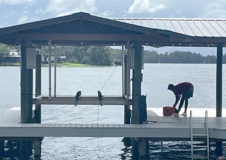 A person in a maroon shirt and shorts works with a red container on a wooden dock under a covered boat lift by a lake.