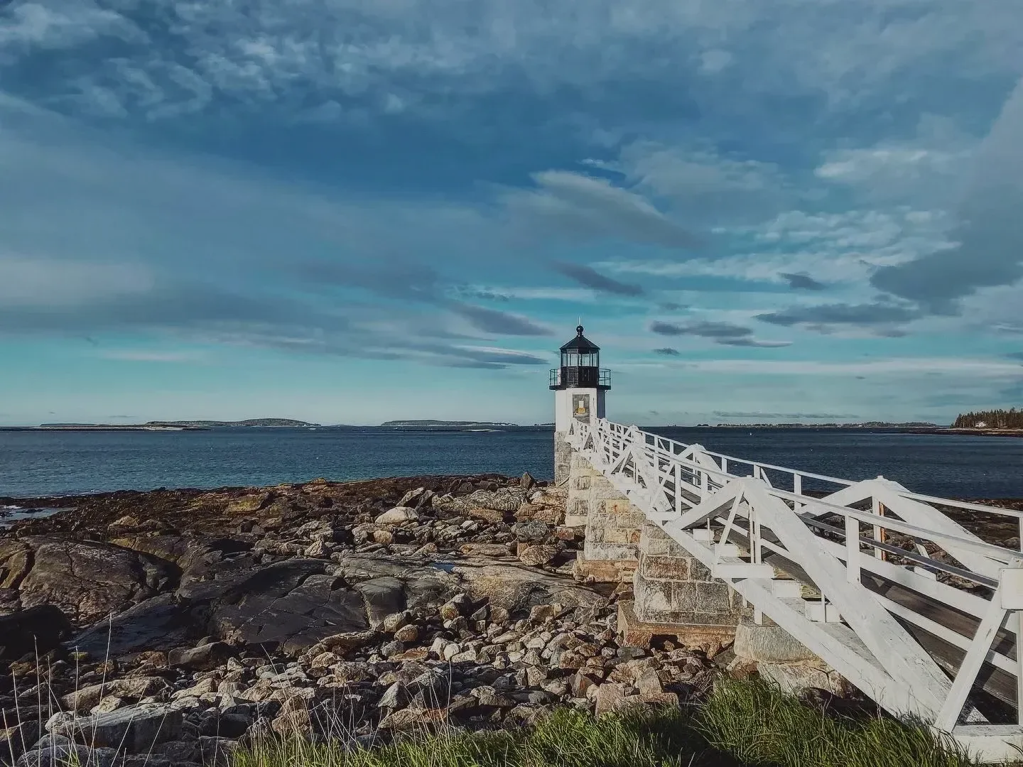 Lighthouse on a rocky shore with a white fence and blue ocean under a cloudy sky