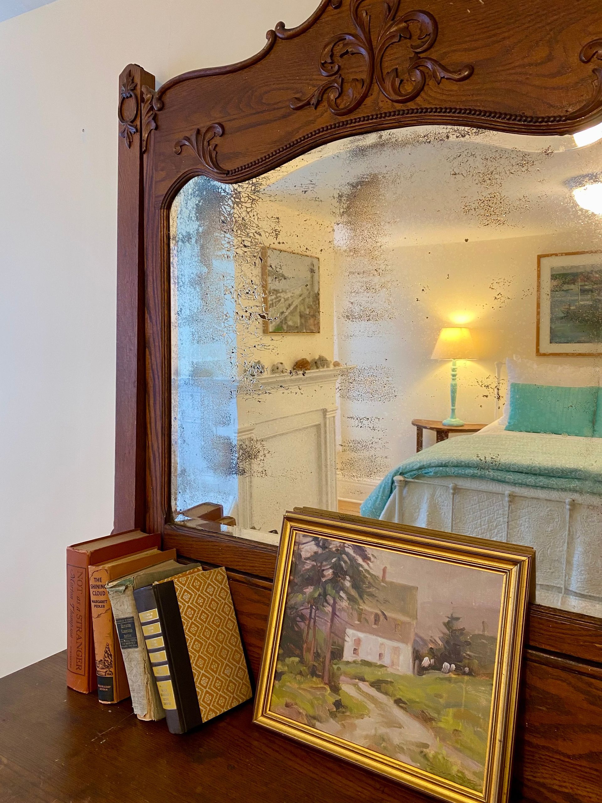 Ornate wooden mirror above framed photos on a dresser in a bedroom, reflecting a bed and lamp.