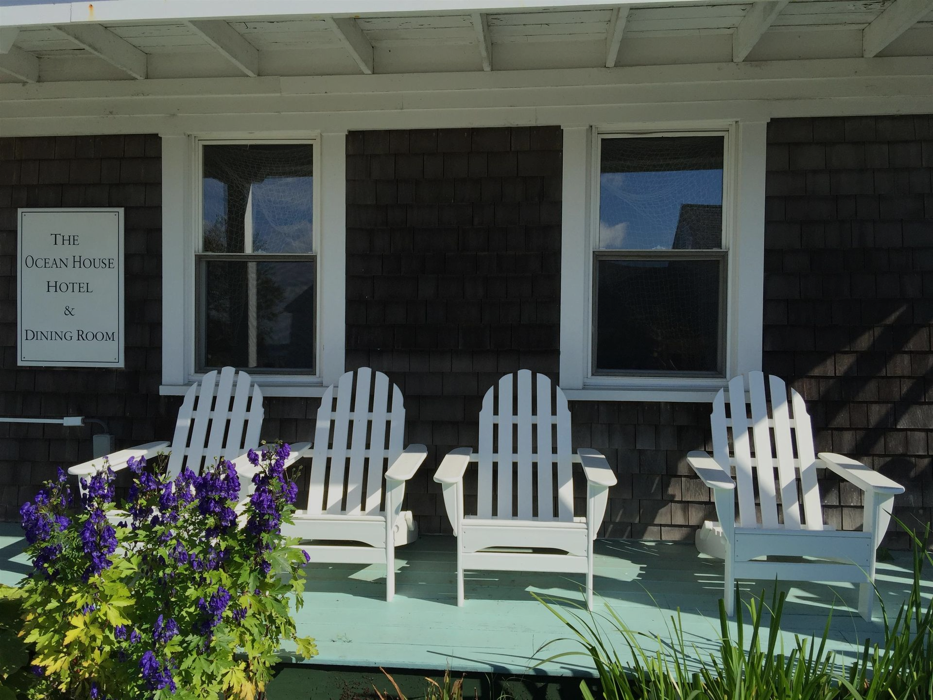 White Adirondack chairs on a porch beside a sign and purple flowers in front of a dark shingled building