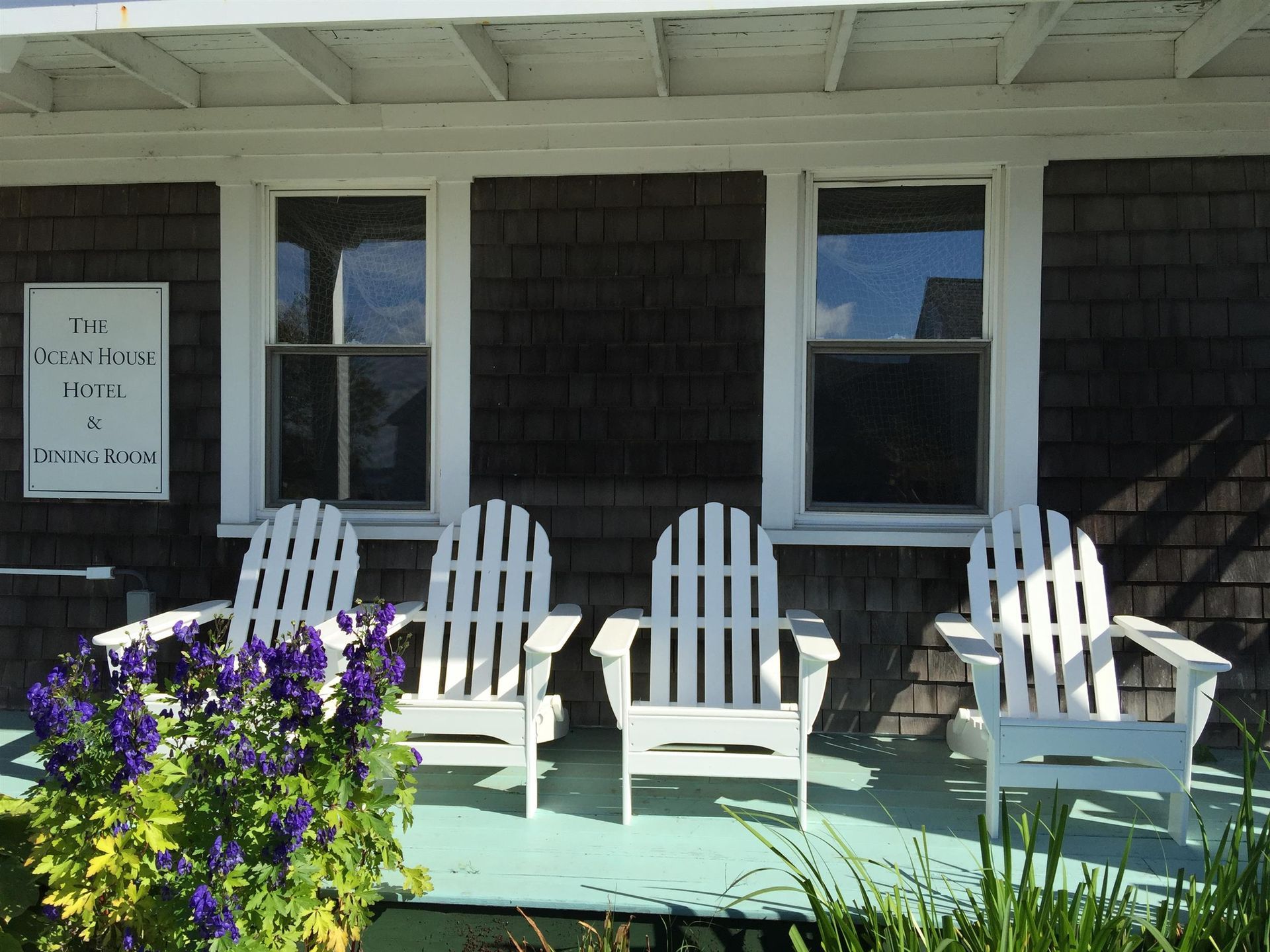 White rocking chairs on a porch in front of a dark-shingled house with windows and purple flowers.
