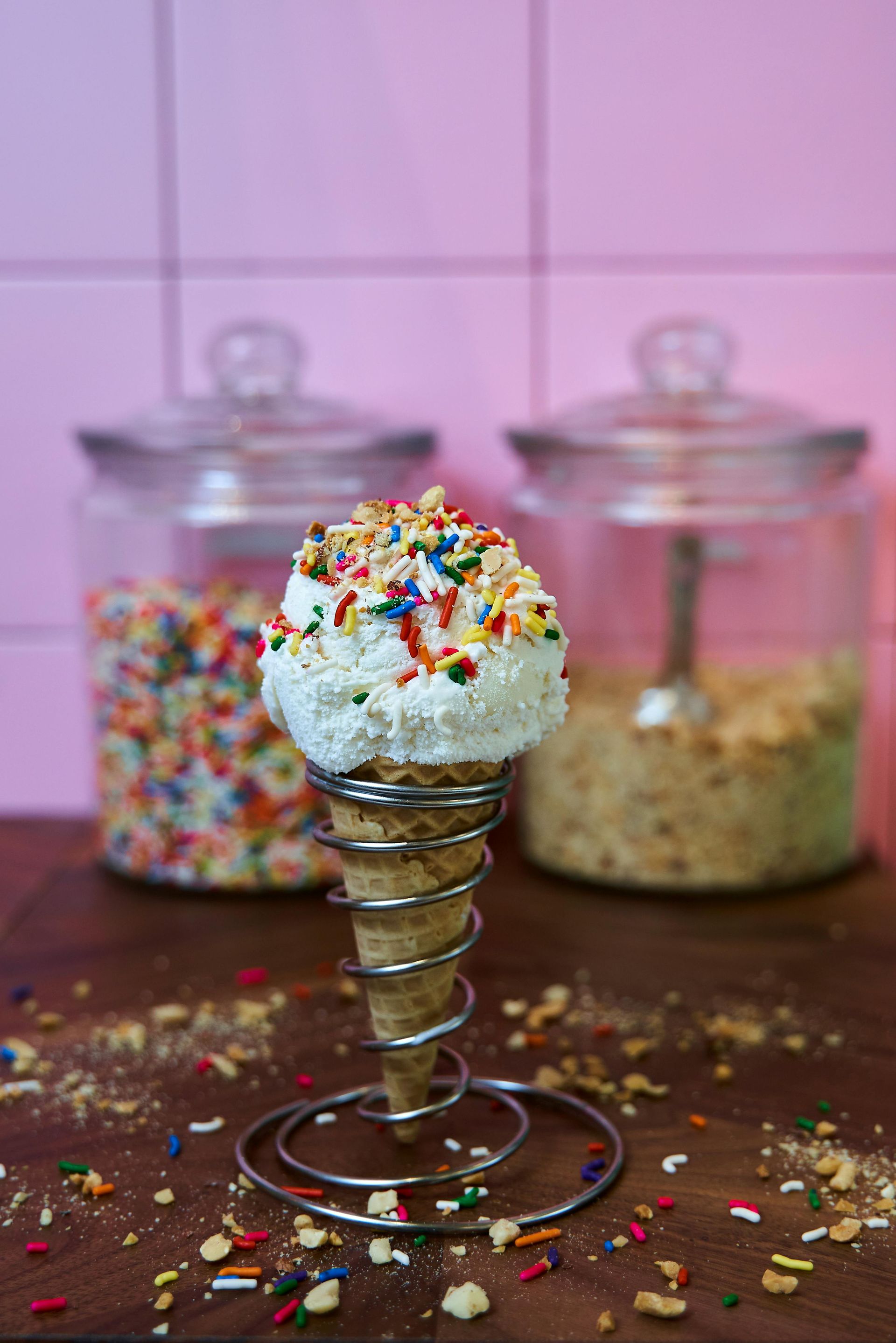 Ice cream cone topped with whipped cream and colorful sprinkles, with candy jars in the background.