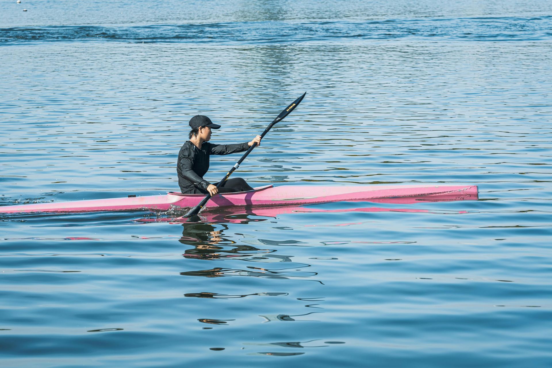 Person kayaking on calm blue water in a pink kayak with a black paddle