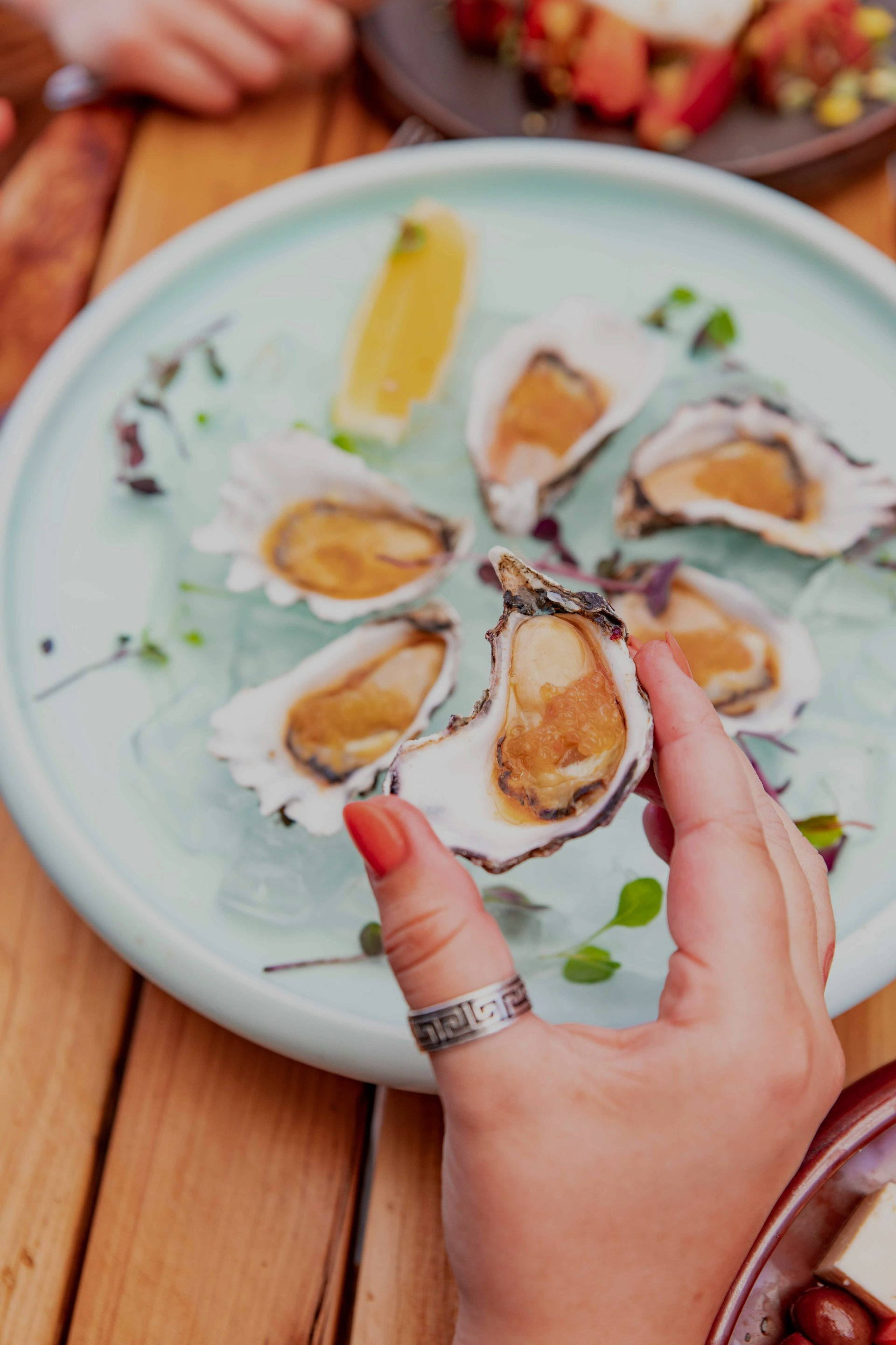Hand holding oyster next to plate of oysters, lemon wedge. Wooden table.