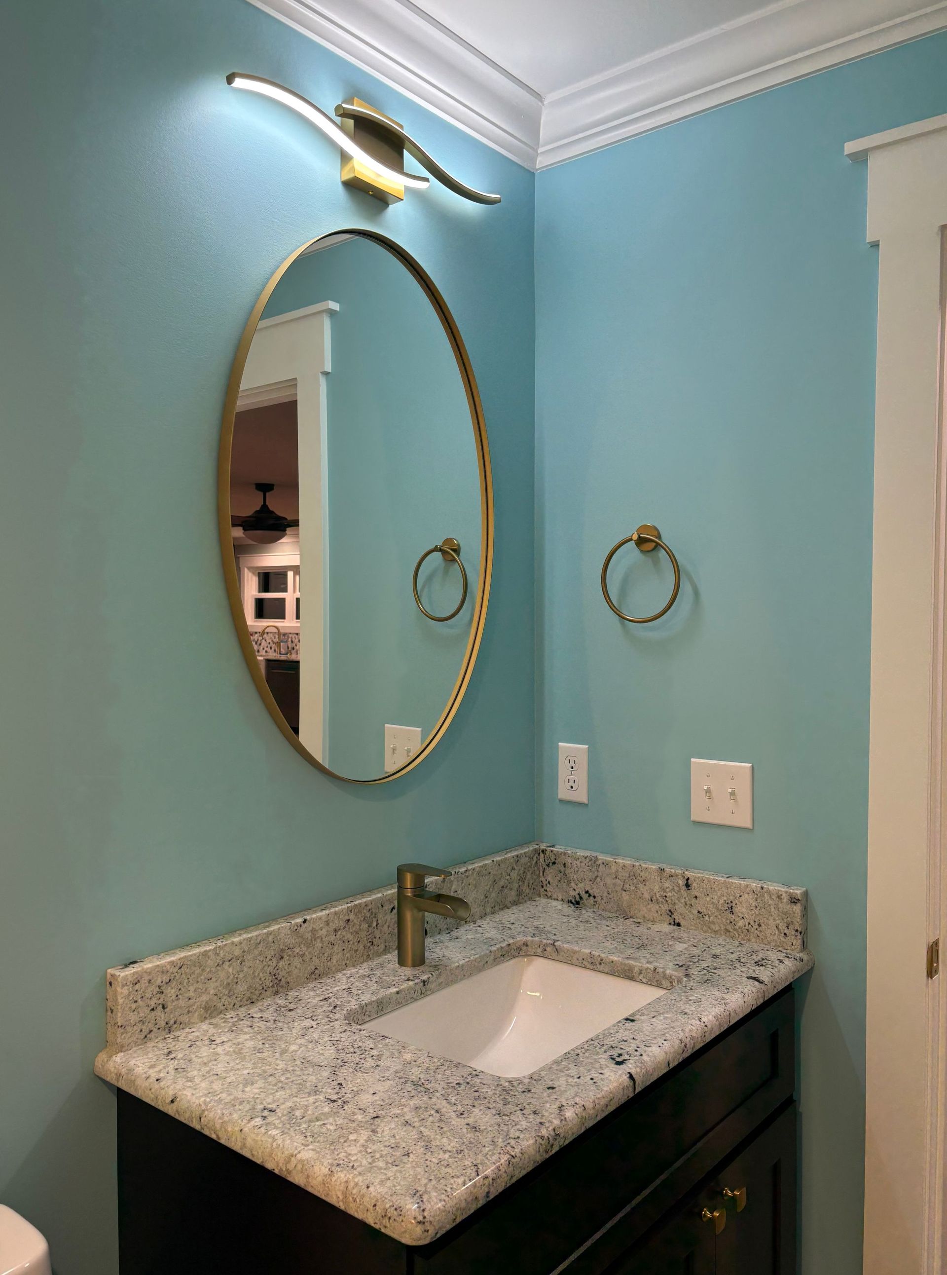Bathroom with blue walls, oval mirror, gold fixtures, granite countertop, and white sink.