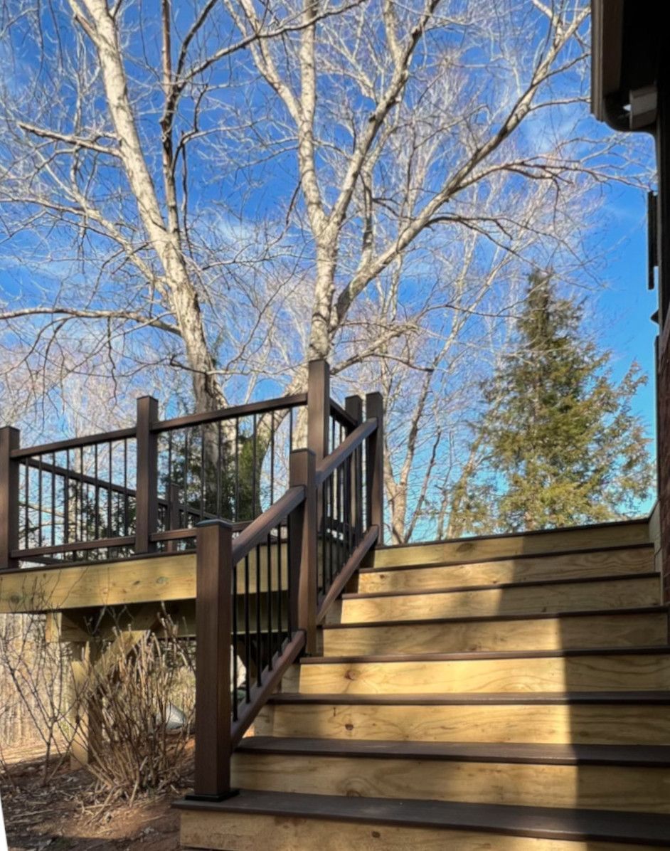 Wooden deck and stairs leading up to it, with railings. Large tree in background, blue sky.