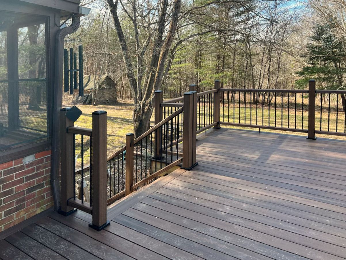 A brown composite deck with railings overlooking a wooded area. Brick house on the left.