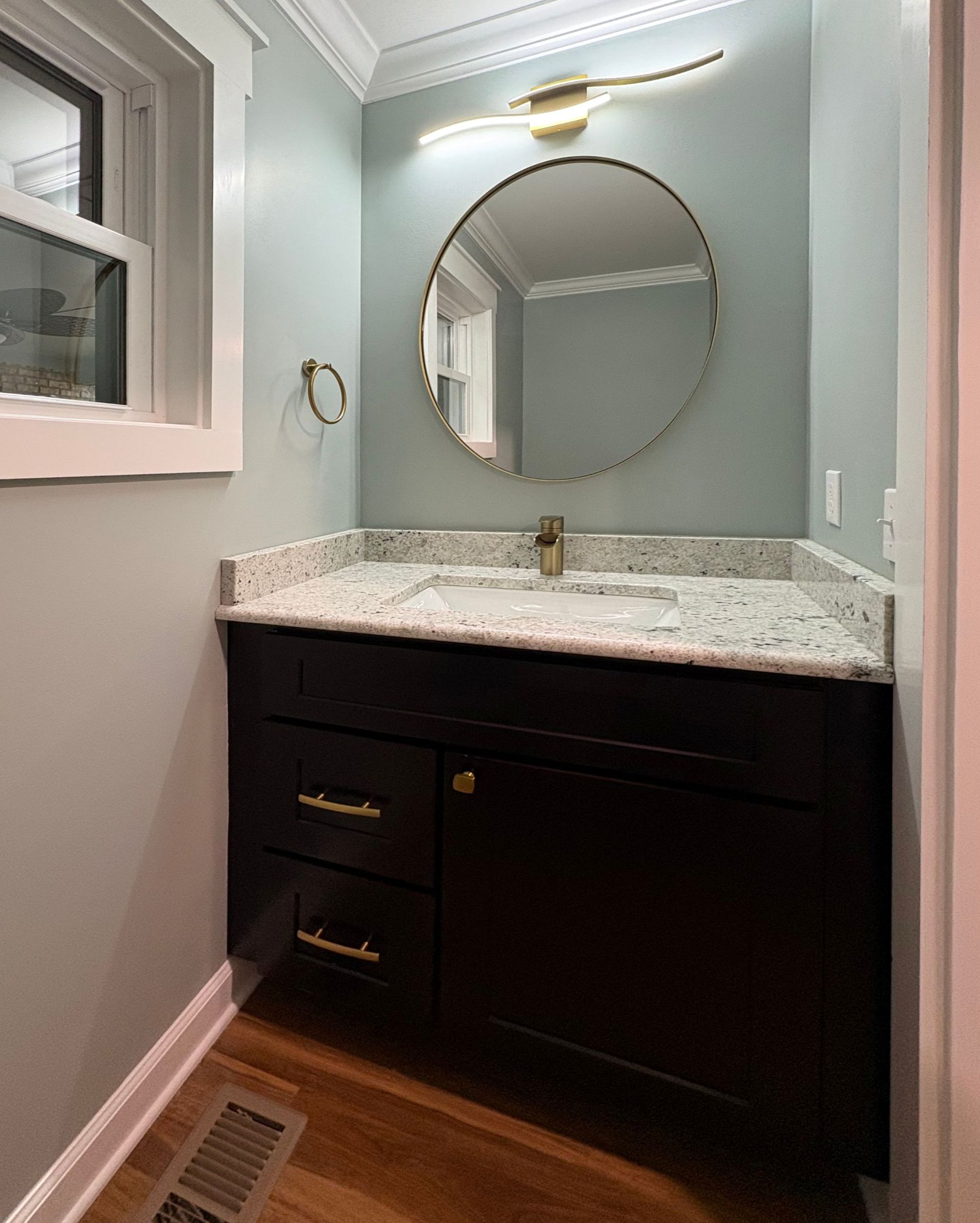 Bathroom with a dark vanity, round mirror, gold fixtures, and light blue walls.