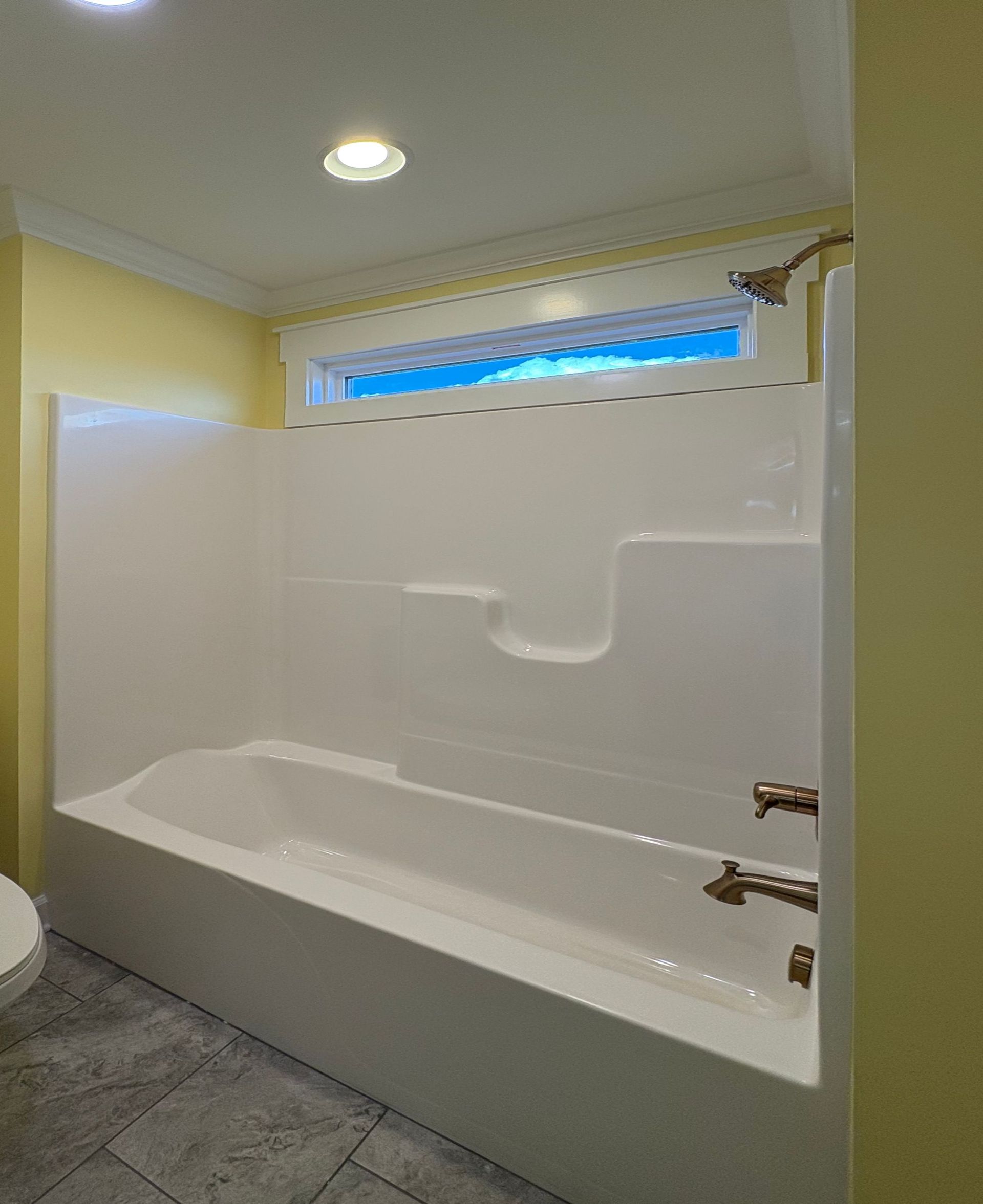 White bathtub and shower surround with window, yellow walls, and gray floor.