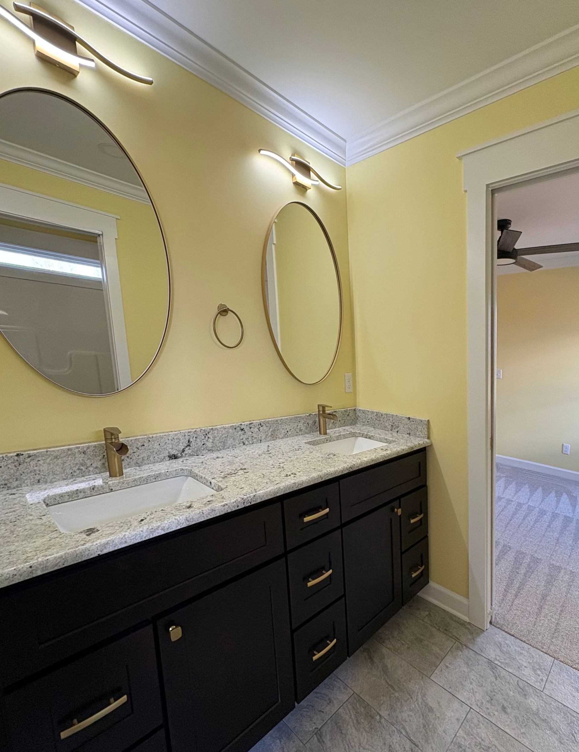 Bathroom with black vanity, granite countertop, gold fixtures, oval and circular mirrors, and yellow walls.