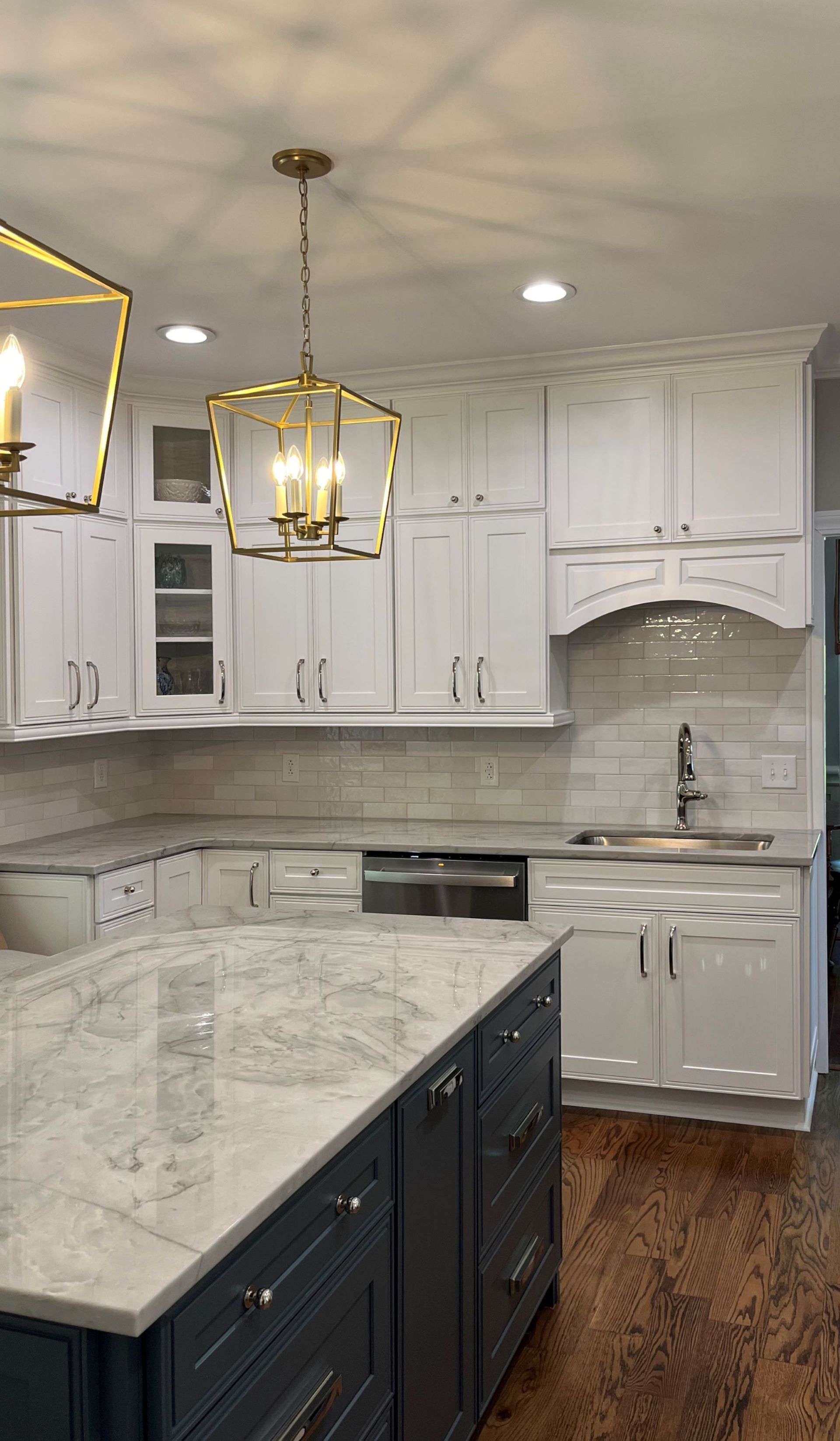White kitchen with dark blue island. Gold light fixtures, white cabinets, and light gray backsplash.
