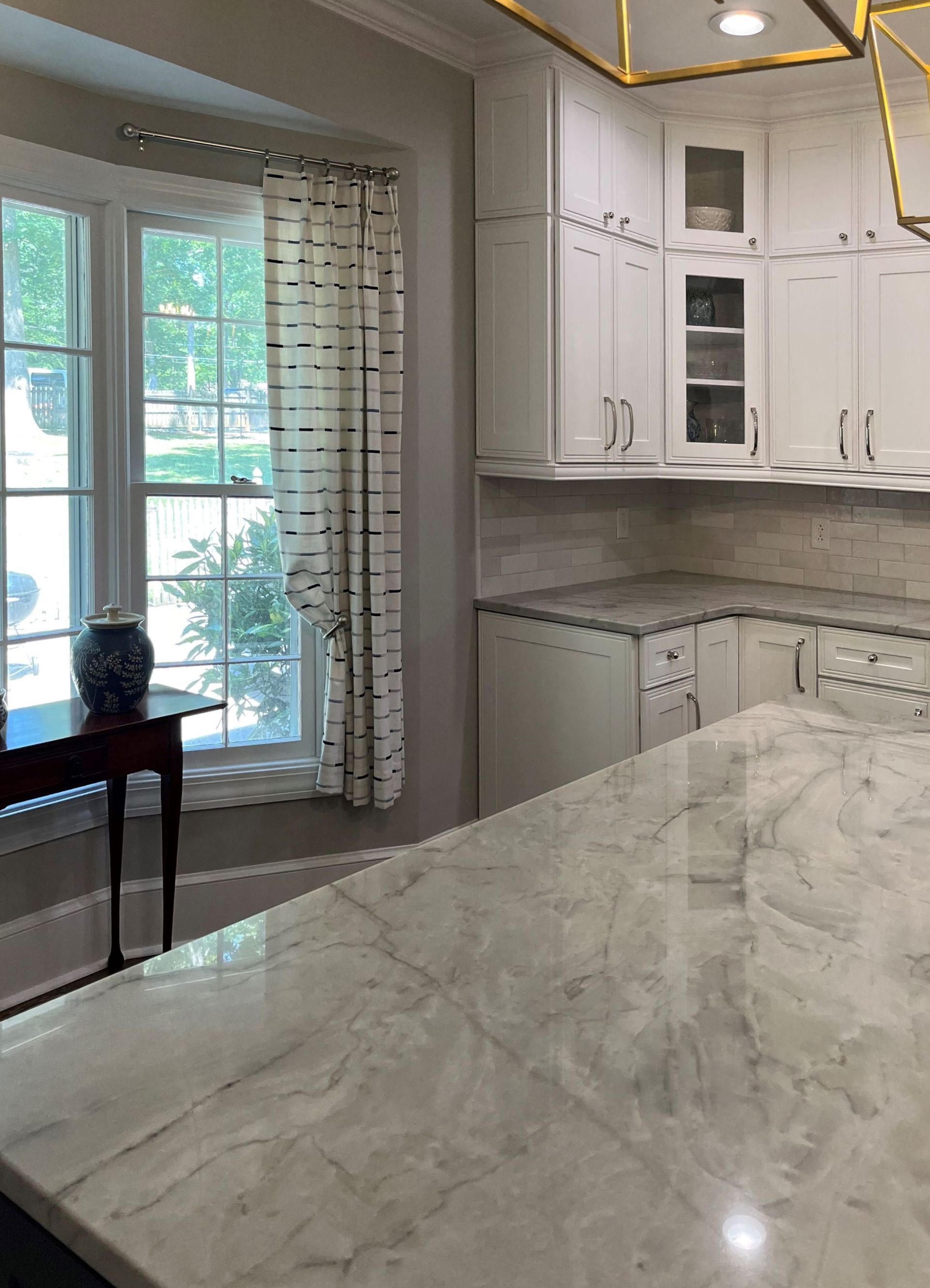 White kitchen with marble countertops, cabinets, and a window with a curtain.