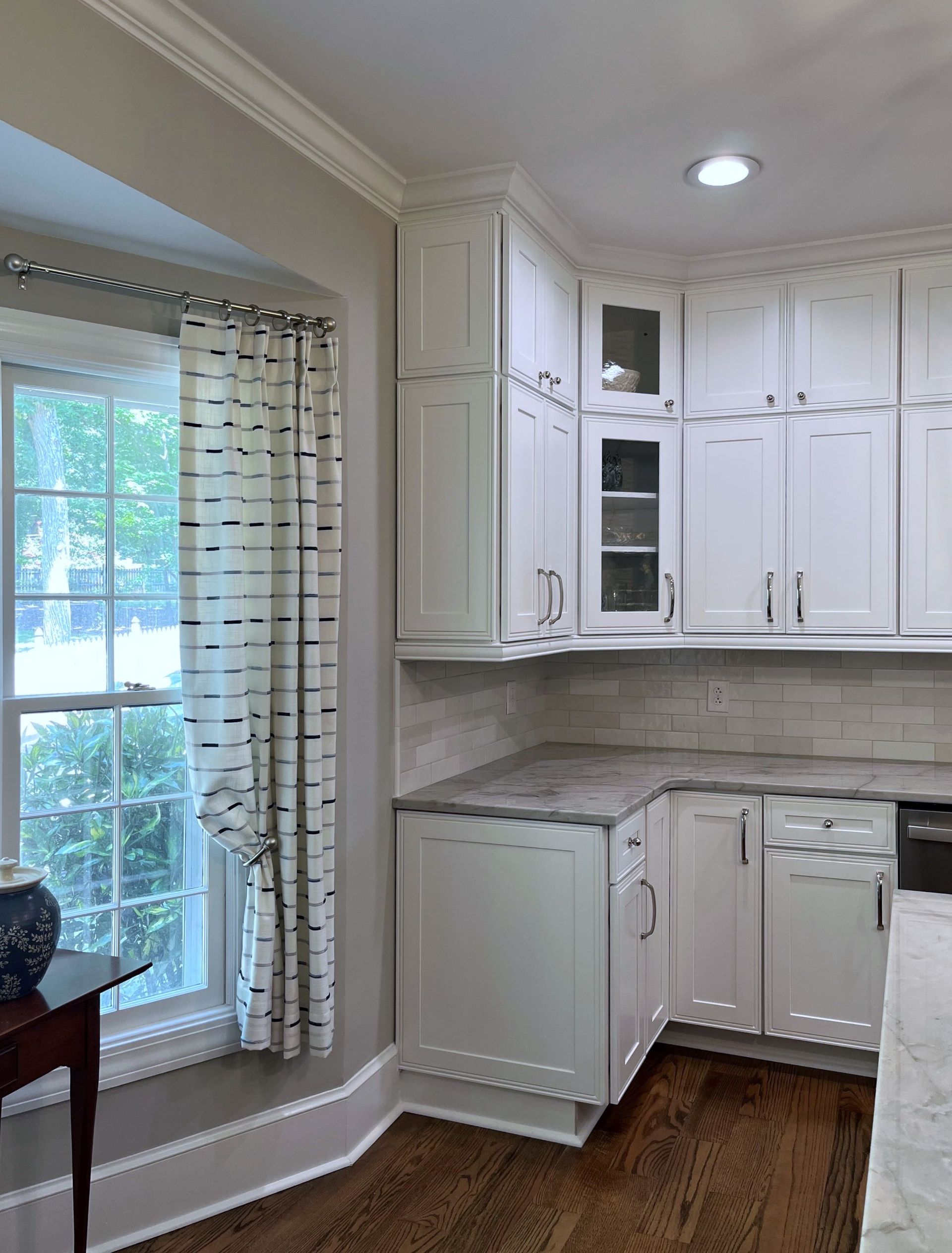 White kitchen cabinets and countertops in a corner, next to a window with patterned curtains.