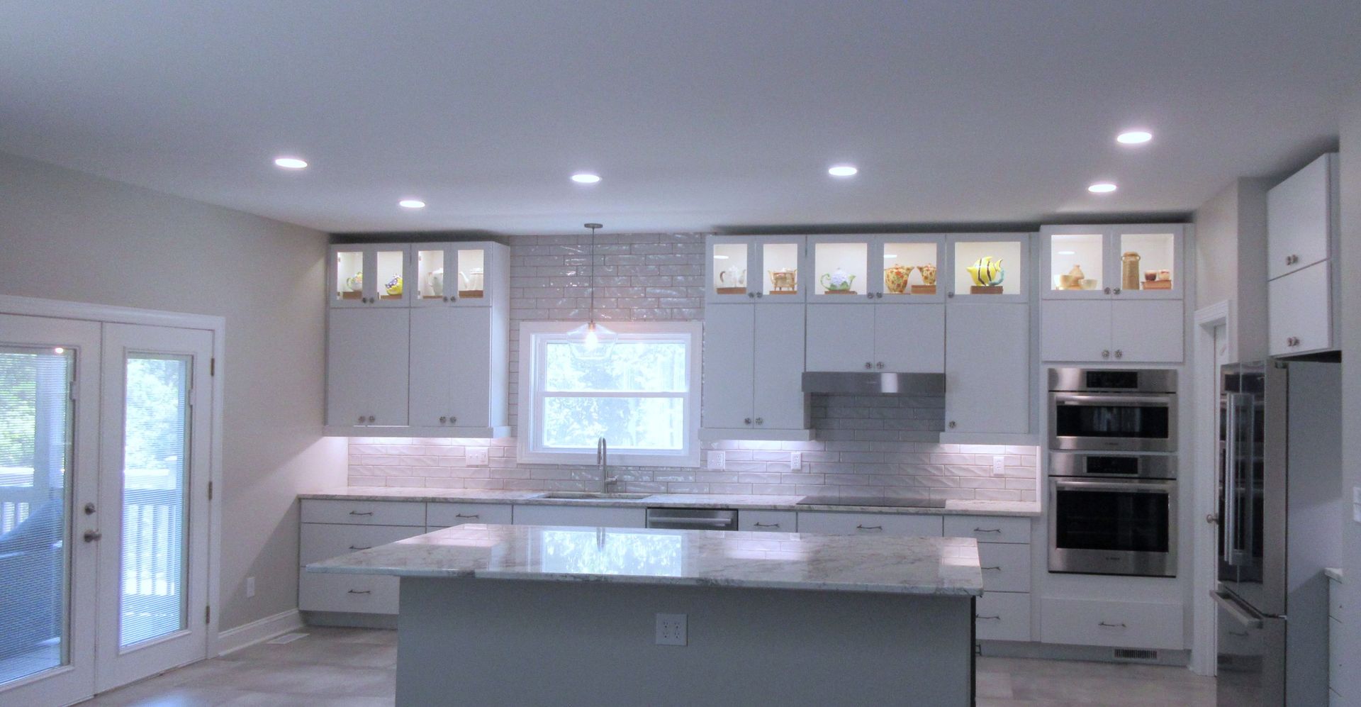 Spacious, modern kitchen with white cabinetry, island, and double ovens. Features natural light from windows and recessed lighting.