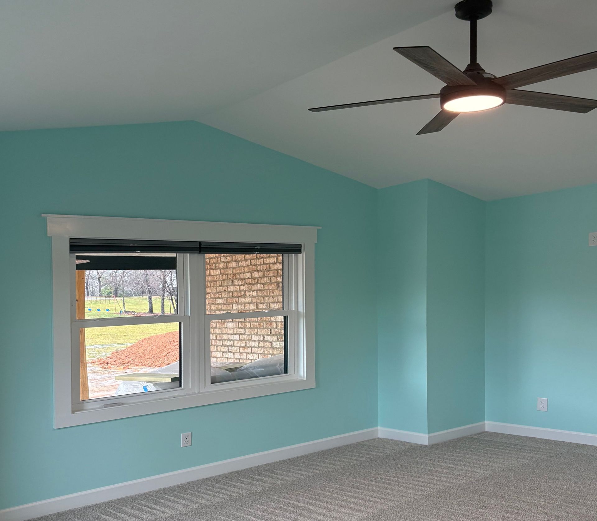 A room with light blue walls, a white-framed window, and a ceiling fan. Grey carpet.