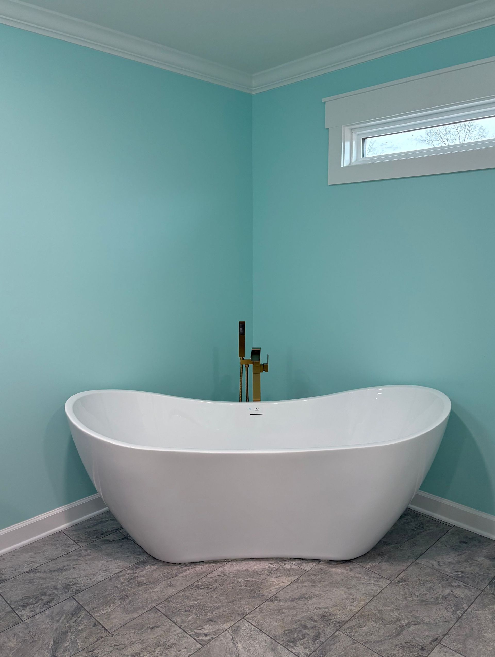 Freestanding white bathtub in corner of aqua-colored bathroom with gold faucet.