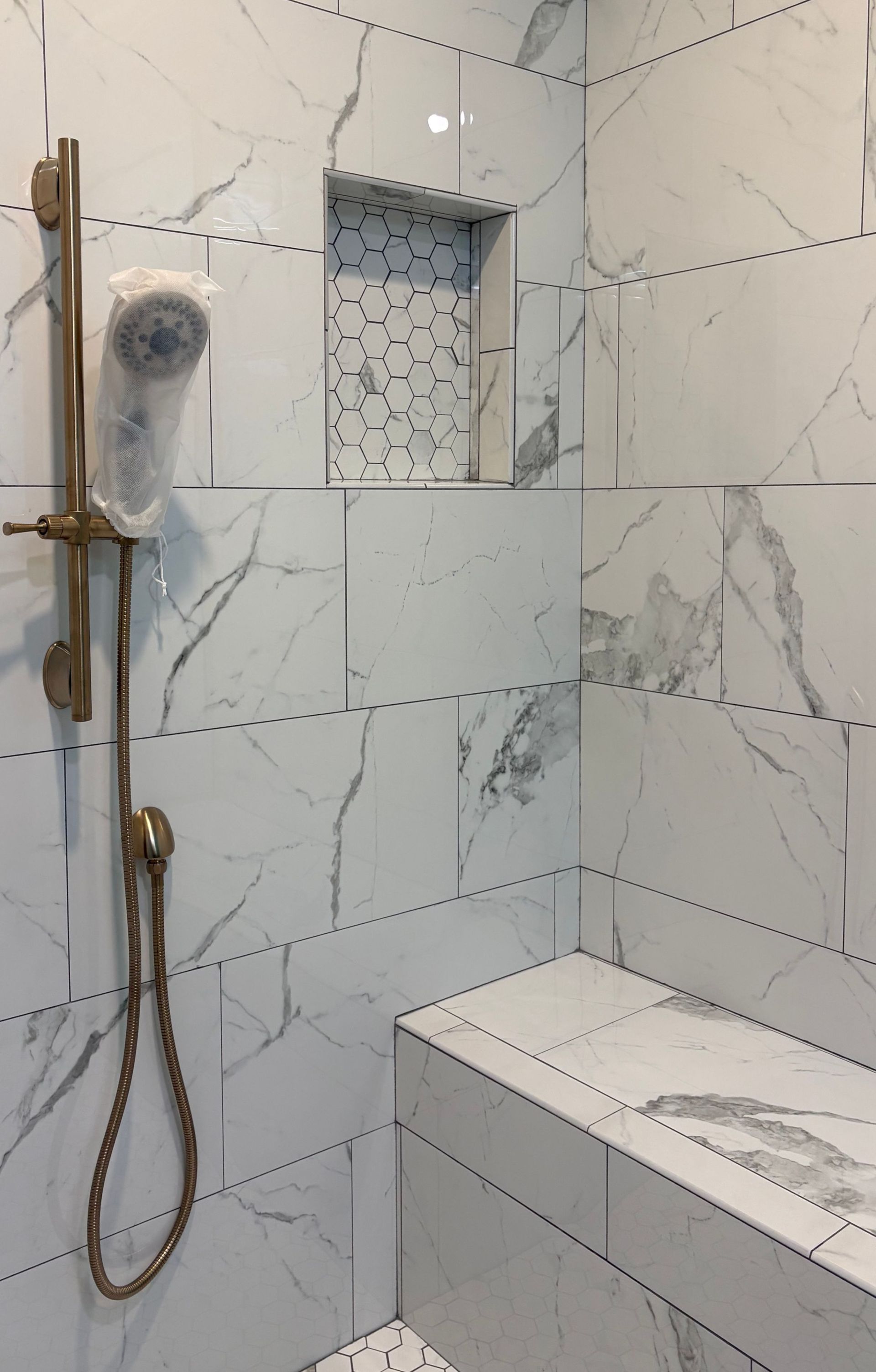 White marble-tiled shower with a gold showerhead and bench. A tiled inset holds shampoo.