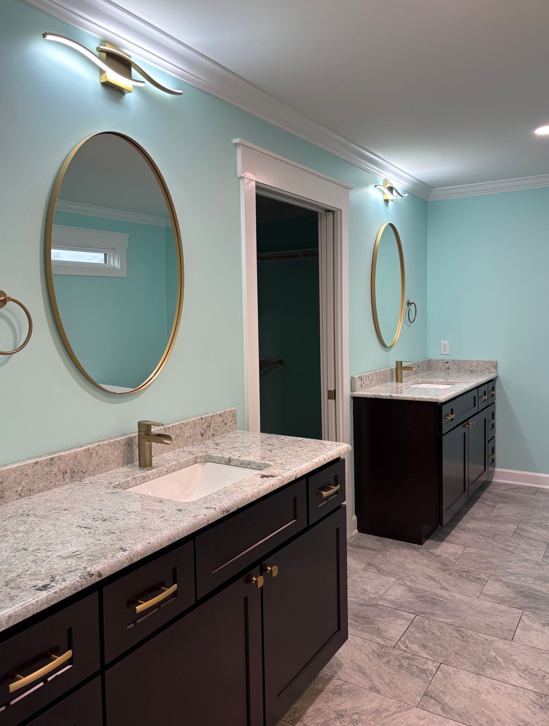 A modern bathroom with two dark cabinets, oval mirrors, and light blue walls.