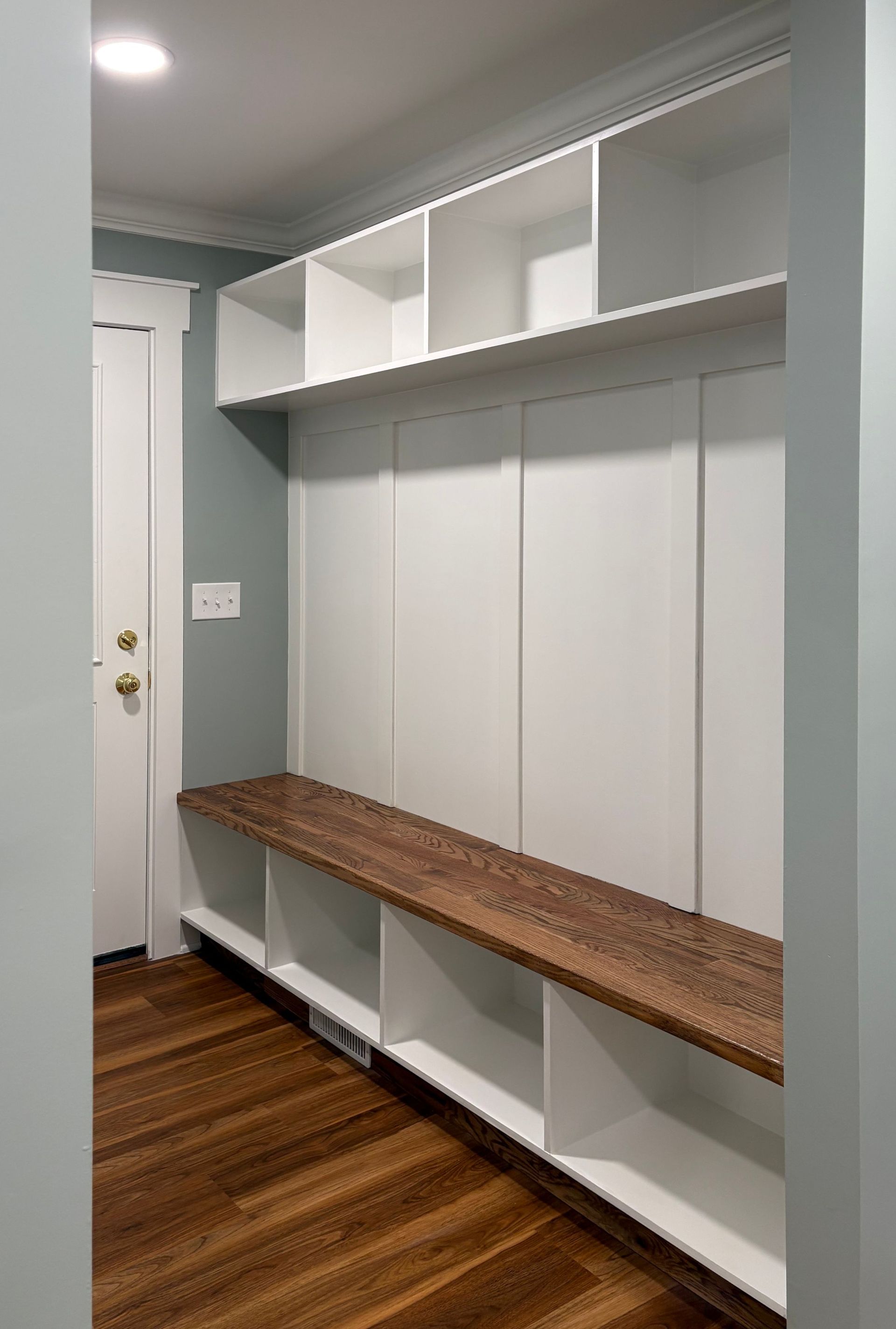 White built-in entryway bench with open storage, a dark wood seat, and upper shelving in a room with a wood floor.