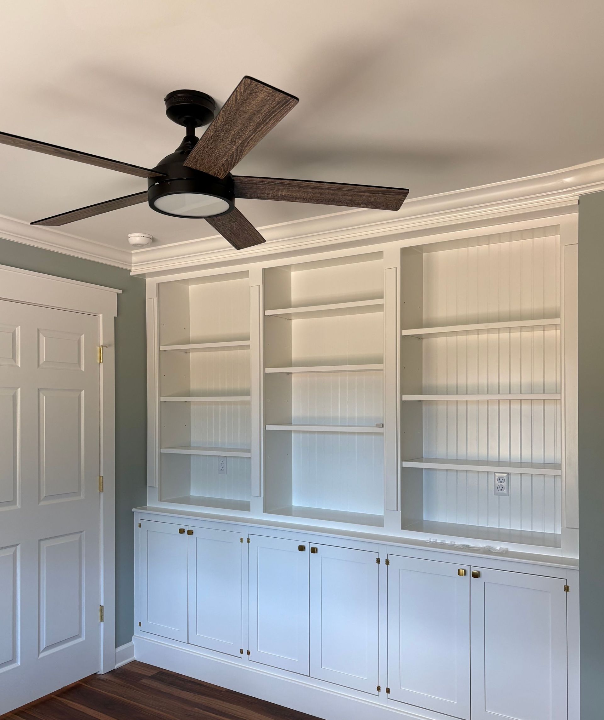 Built-in white bookshelves with cabinets, door on left, ceiling fan overhead.