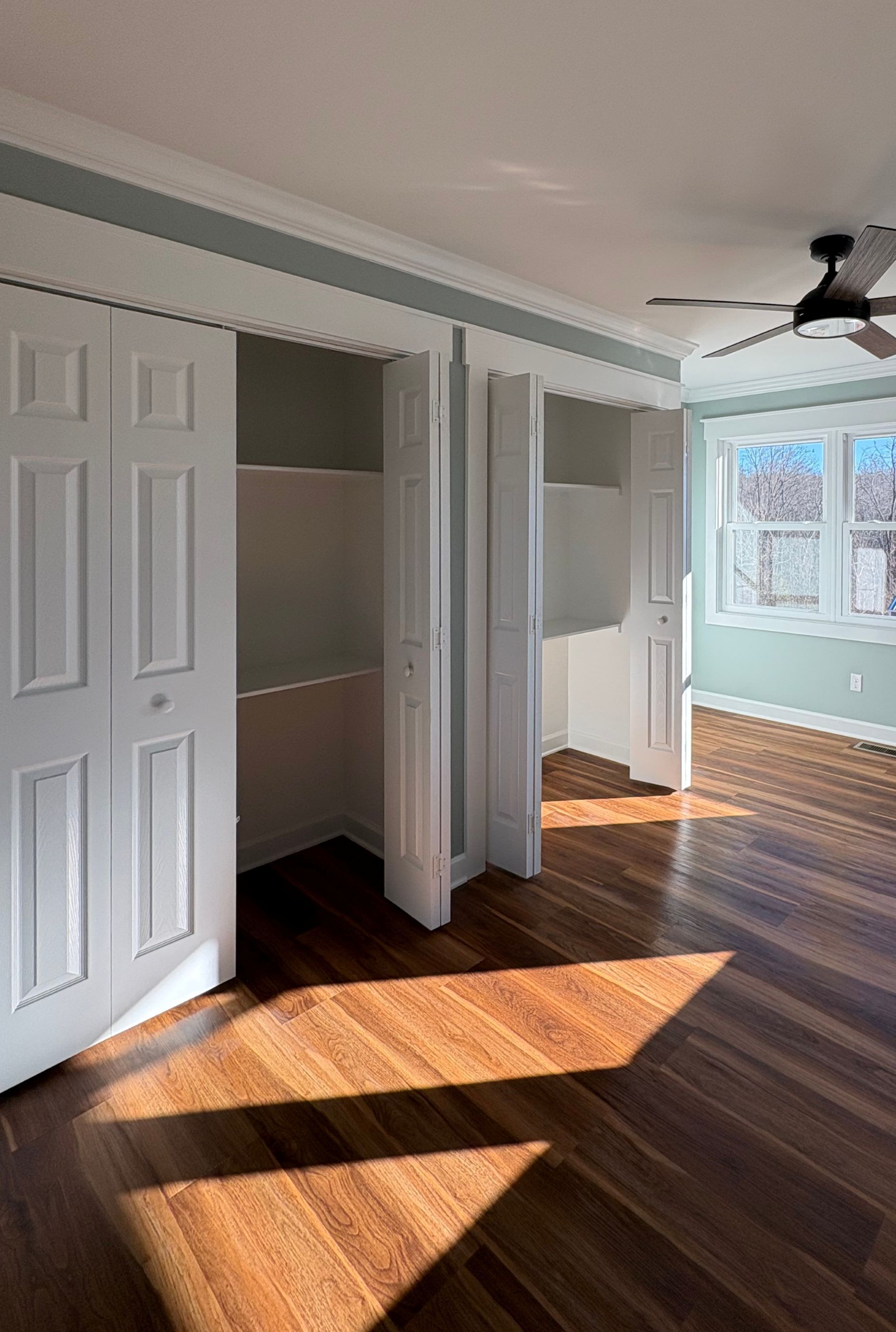 Open white closet doors in a bedroom with wood floors, sunlight, and a window.