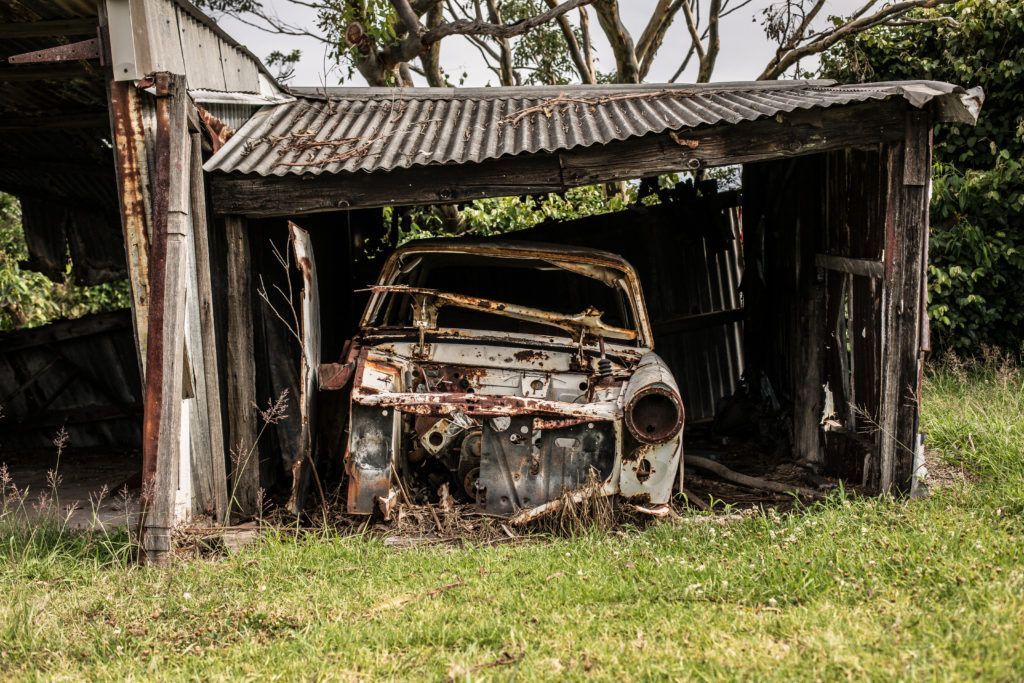 An old rusty car is parked under a wooden shed.