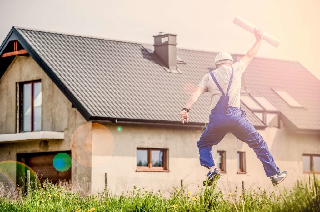 A man in overalls is jumping in the air in front of a house.