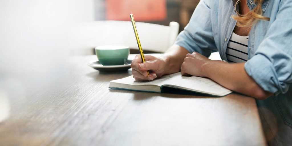 A woman is sitting at a table writing in a notebook with a pencil.