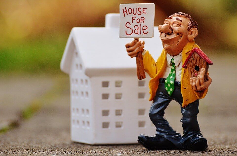 A figurine of a man holding a house for sale sign in front of a small house.