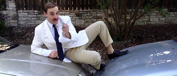 A man in a white suit and tie is sitting on the hood of a car.