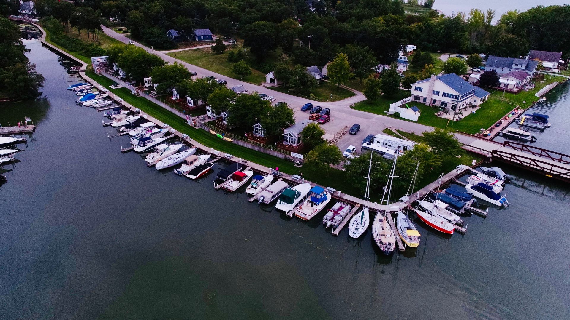 Aerial view of a marina with many docked boats beside a tree-lined waterfront and houses.