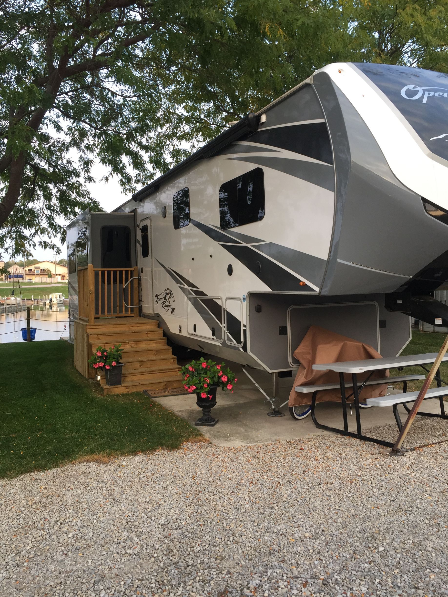 Large gray-and-black RV parked on gravel beside a small wooden deck and flowers under trees.
