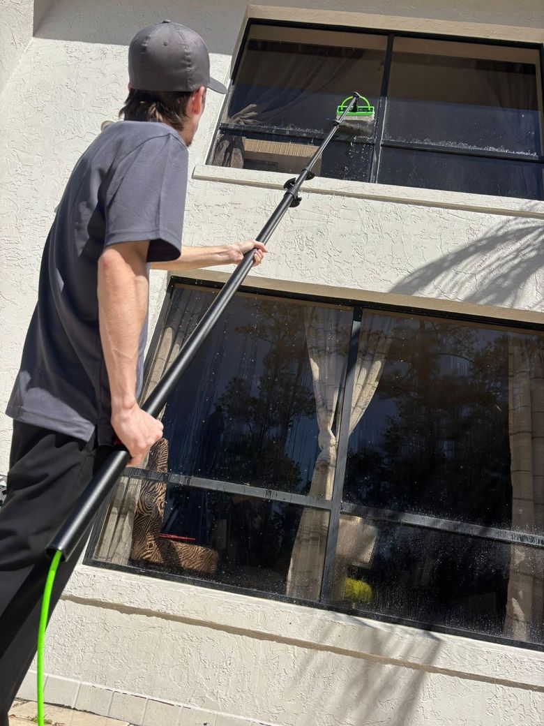 Person using a long-handled cleaning tool to wash an upper window outside a building.