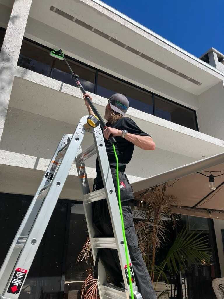 Person on a ladder using a long pole to work on a tall modern building exterior