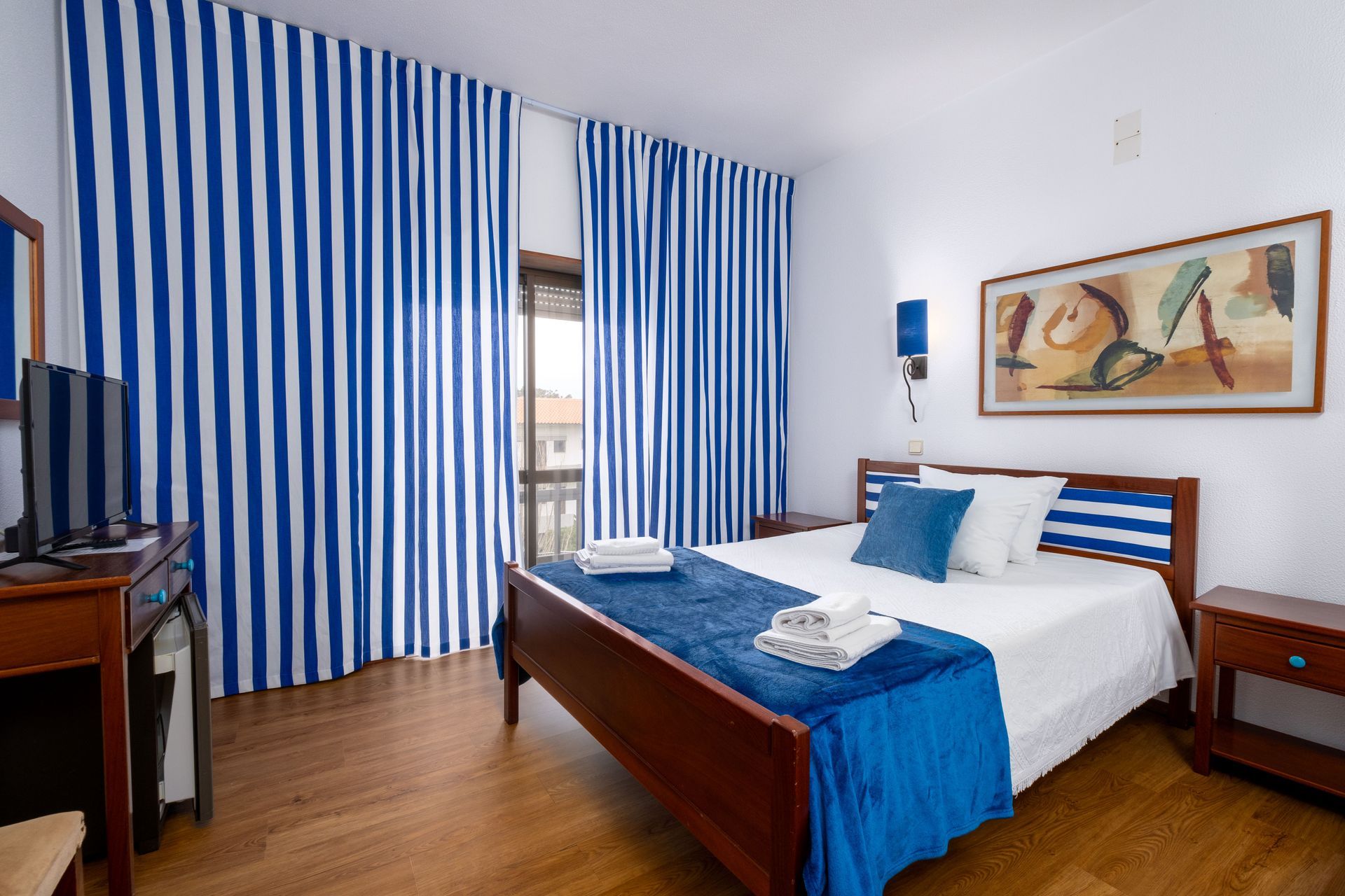 Um quarto com uma cama, mesa de cabeceira, televisão e cortinas azuis e brancas.