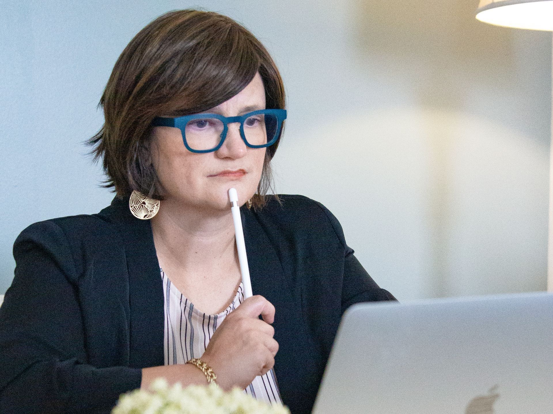 A woman wearing glasses is sitting in front of a laptop computer holding a pen to her mouth.