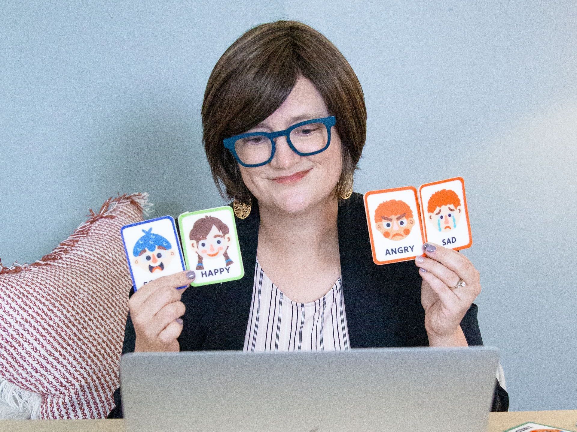 A woman wearing glasses is sitting in front of a laptop holding cards with cartoon faces on them.