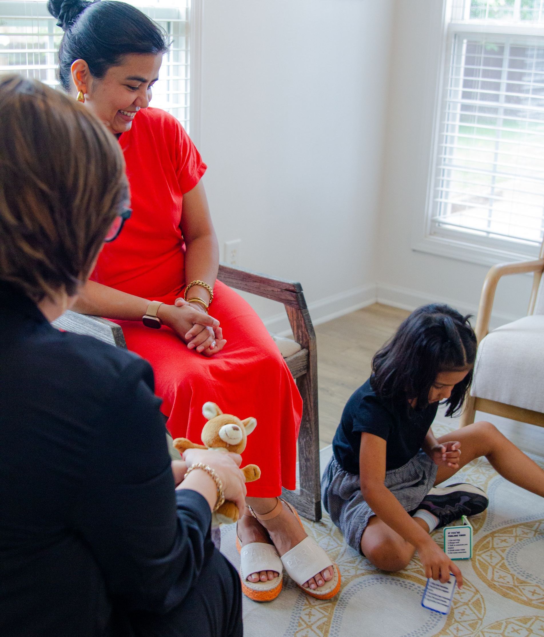 A woman in a red dress is sitting on a chair next to a little girl playing with a teddy bear.