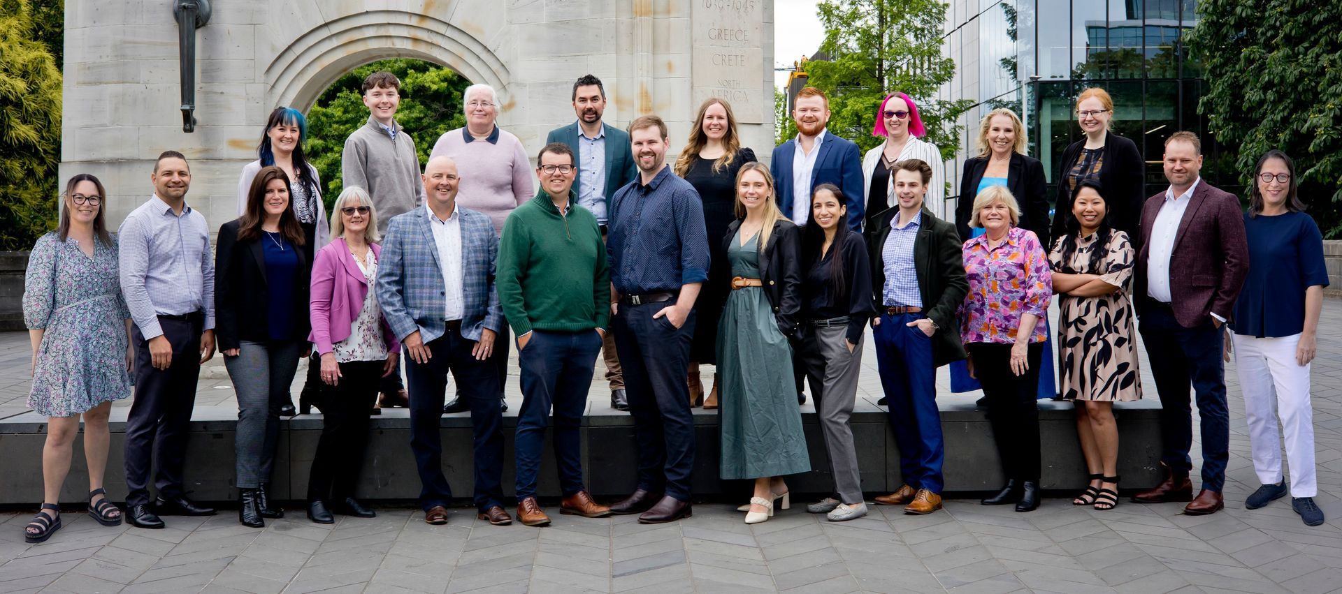 Full team photo of the Rodgers & Co team. Lined up, two rows of people, dressed professionally standing in front of the Bridge of Remembrance in Christchurch. The day is sunny and everyone looks happy. 