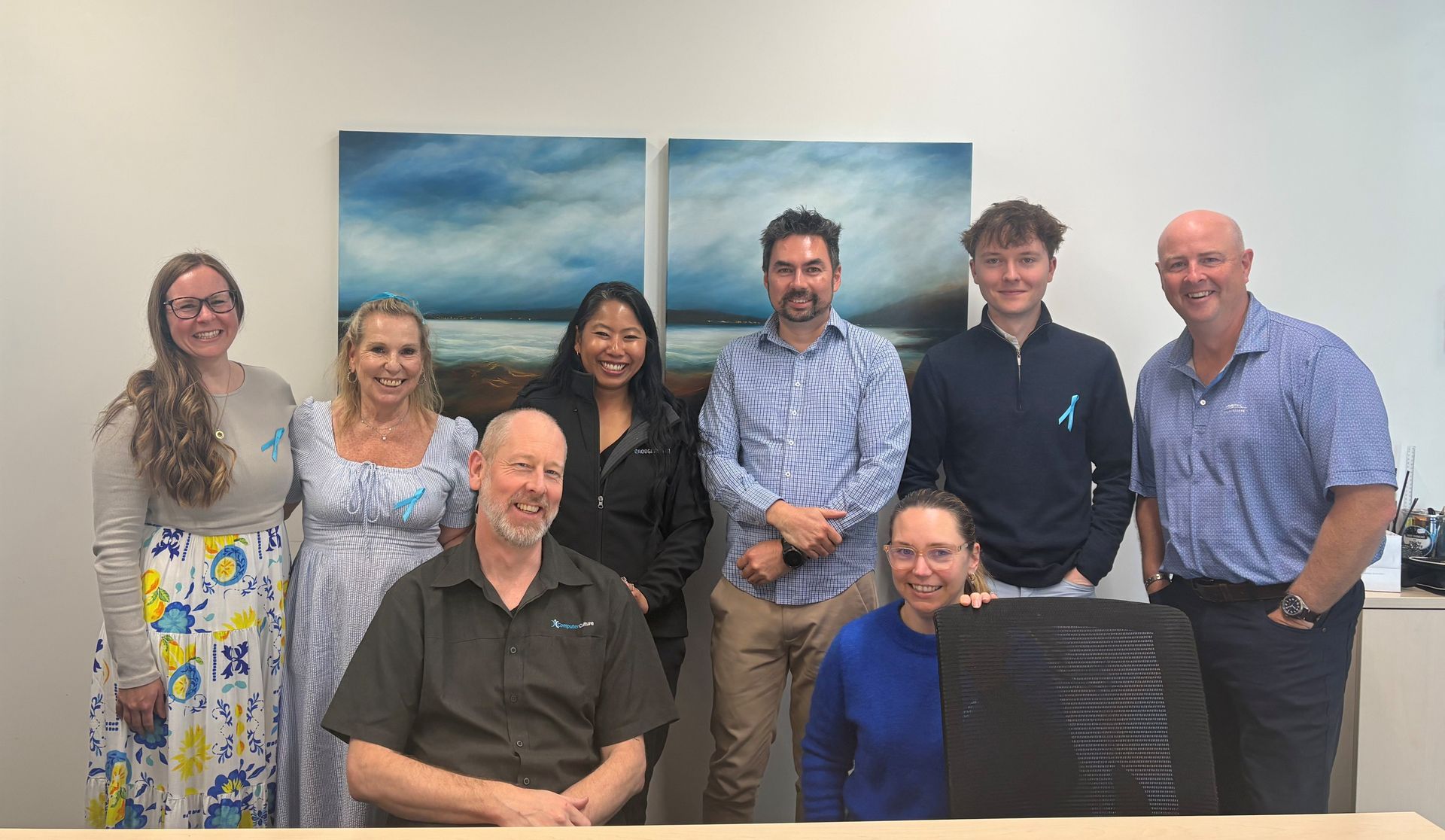 A photo of the Rodgers & Co team in their offices, alongside Tushka Schapel from Computer Culture. The back row has six people standing in front of two landscape paintings, the front row has two people seated (including Tushka from Computer Culture). They are all smiling and happy.