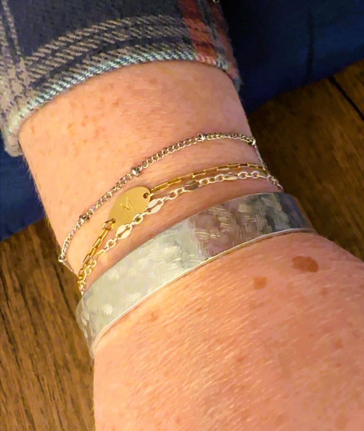 A wrist wearing four bracelets: a silver cuff, a beaded chain, a gold chain with a heart pendant, and a delicate silver chain.