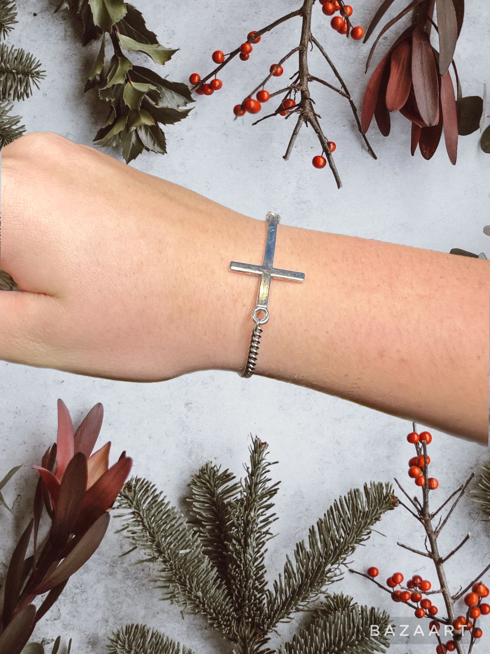 Hand wearing a silver cross bracelet, decorated with berries and greenery on a gray surface.