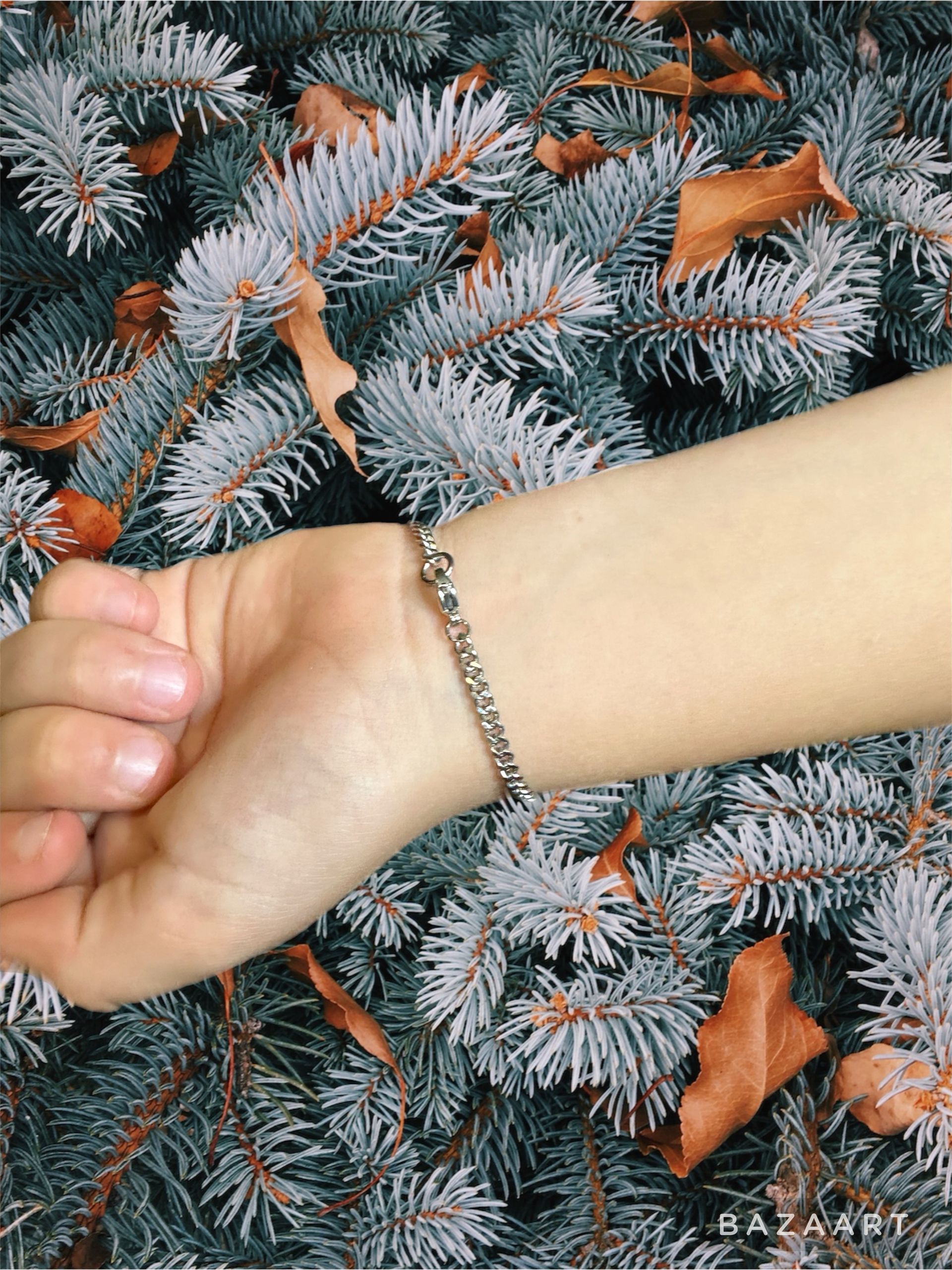Person's wrist with silver chain bracelet, resting on blue-green pine branches with brown leaves.