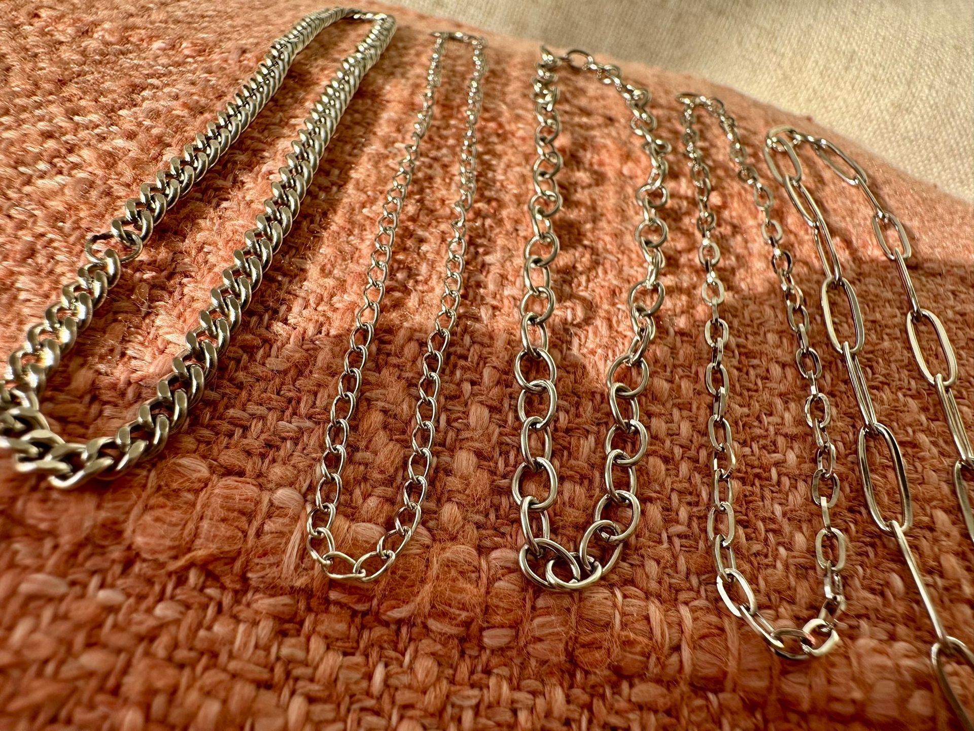 Six silver necklaces of various chain styles on a pink, textured surface.