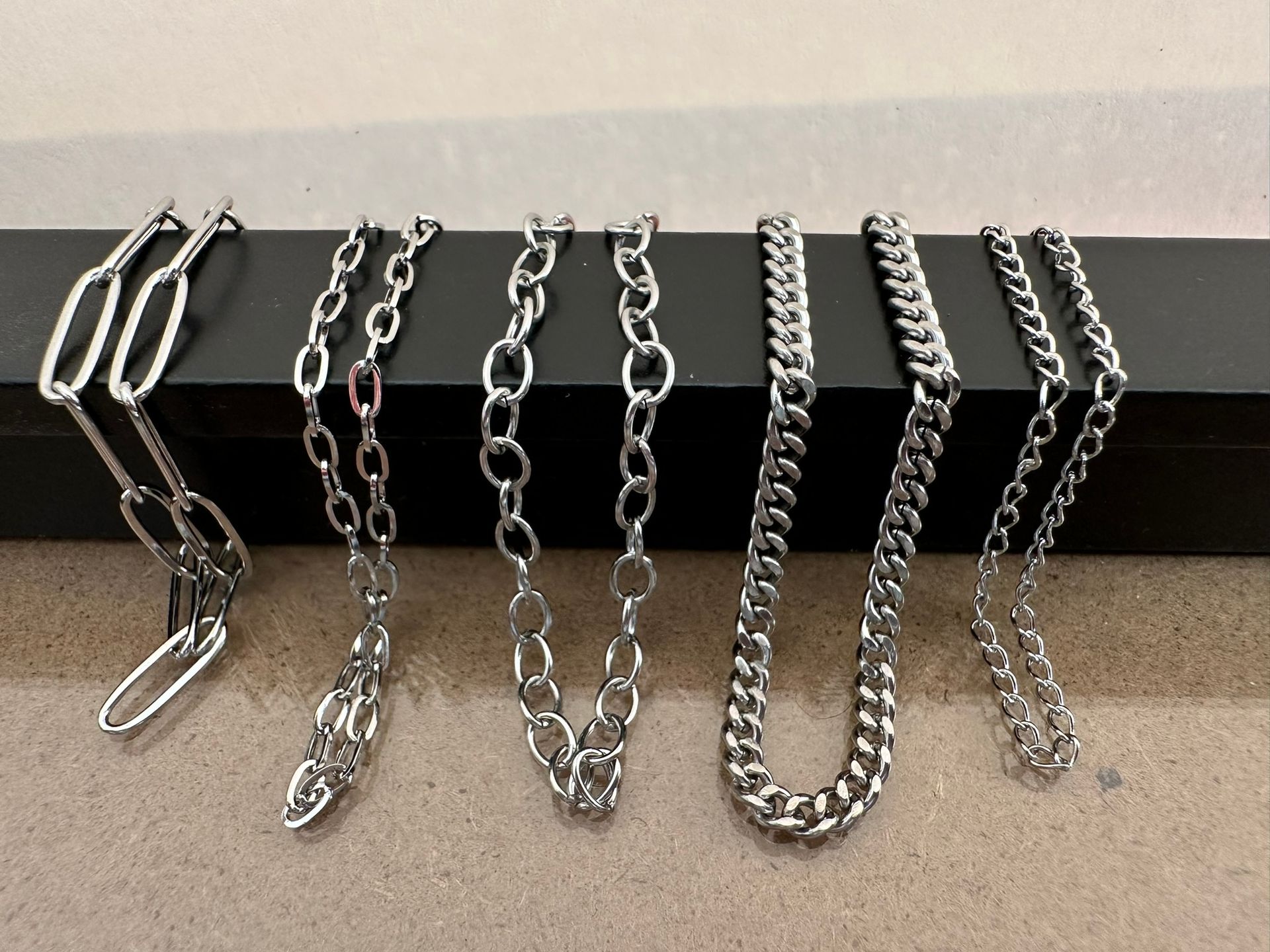 Six silver necklaces displayed on a black surface against a white wall. Different chain styles are visible.