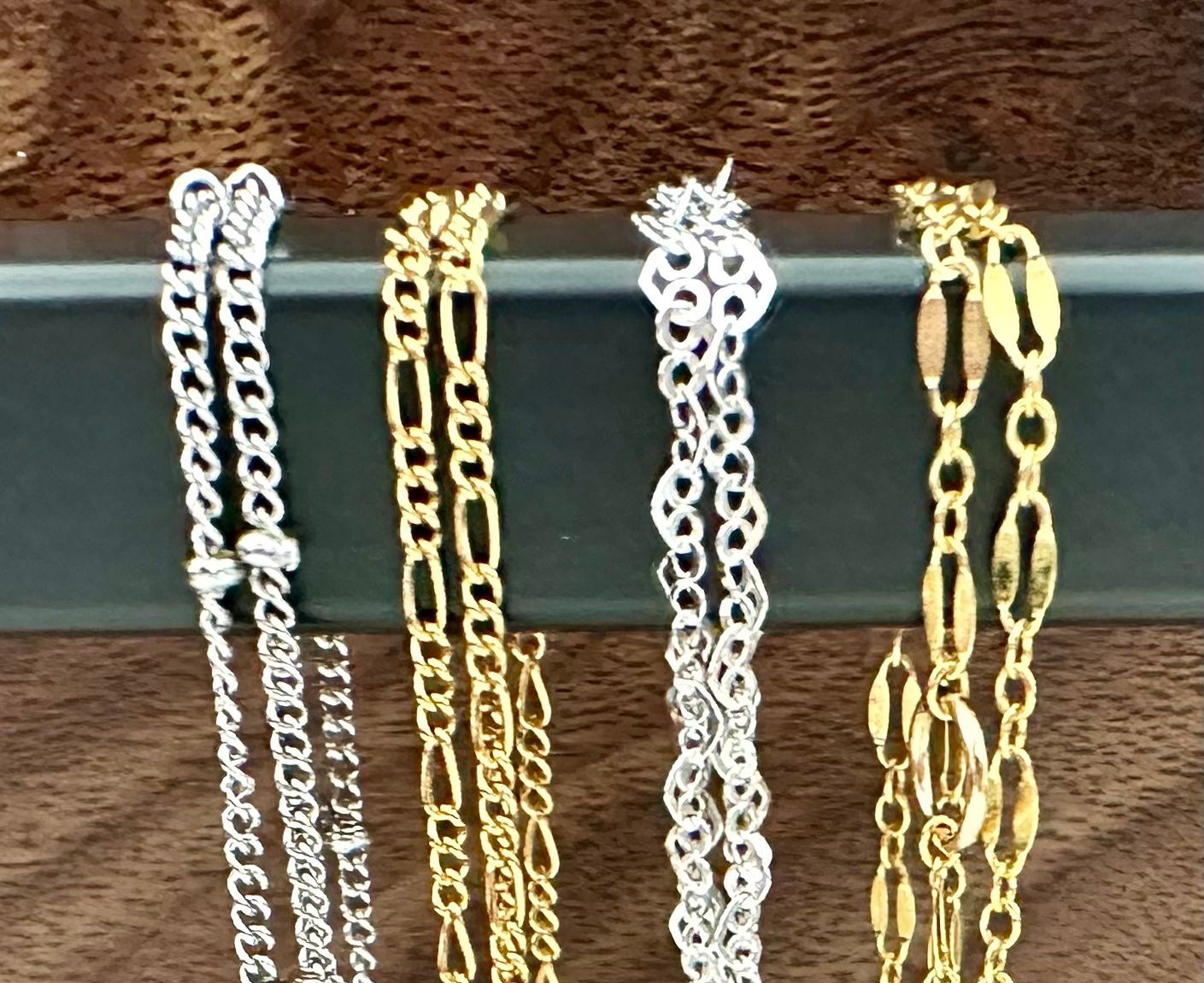 Four gold and silver necklaces hanging on a dark bar, showcasing different chain styles.